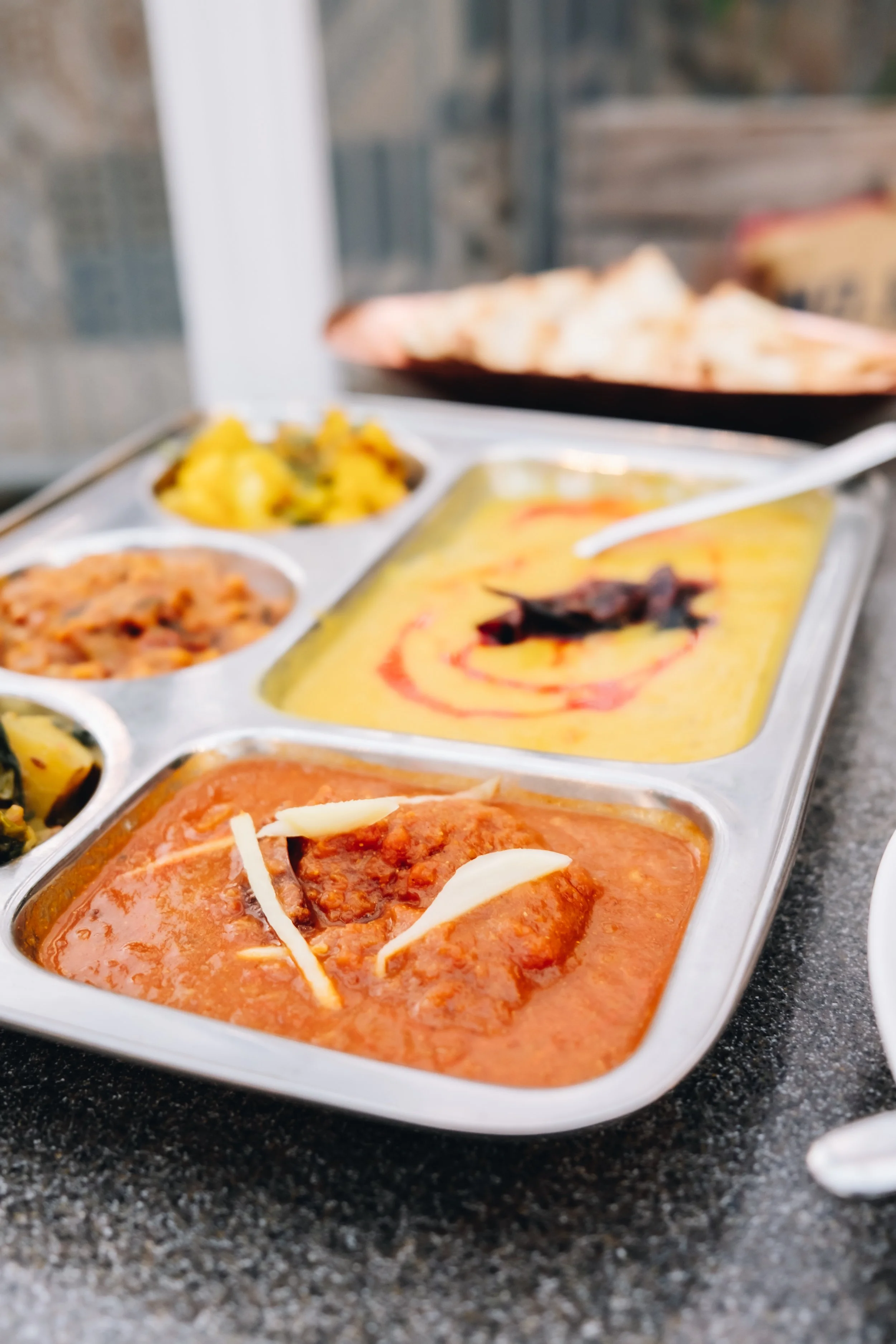 Indian dishes served in a stainless steel tray, including curry with cheese garnish, yellow lentil dal, and other vegetable dishes, with naan bread in the background.