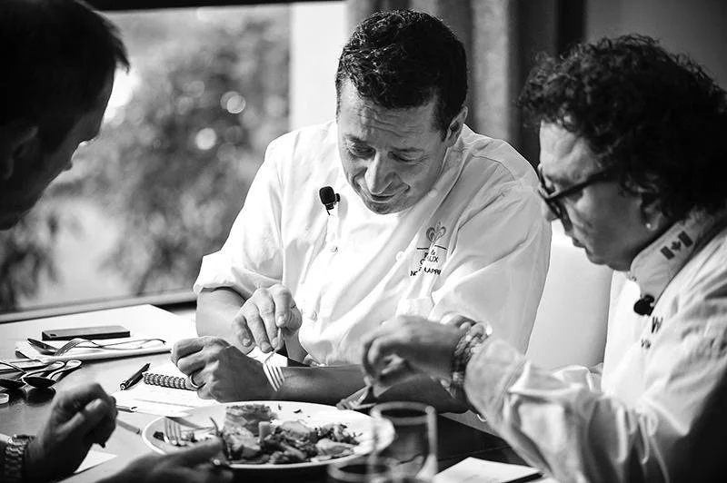 Three men in white chef coats eating together at a table, with utensils and food on the table, in a black-and-white photo.