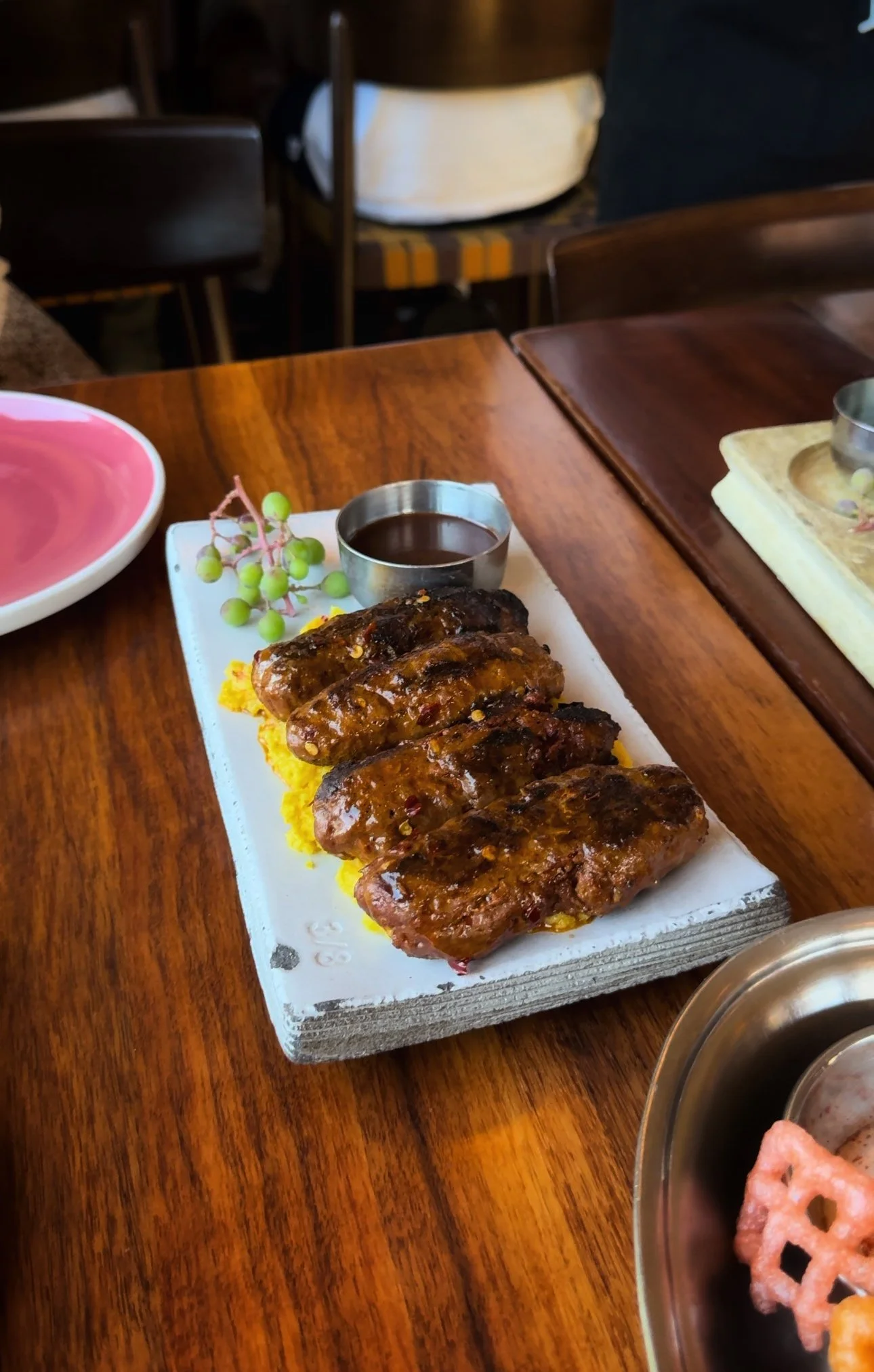 Plate with grilled sausages on a bed of yellow rice, garnished with a small cluster of green grapes, and a small container of dark sauce on a rectangular ceramic plate on a wooden table.