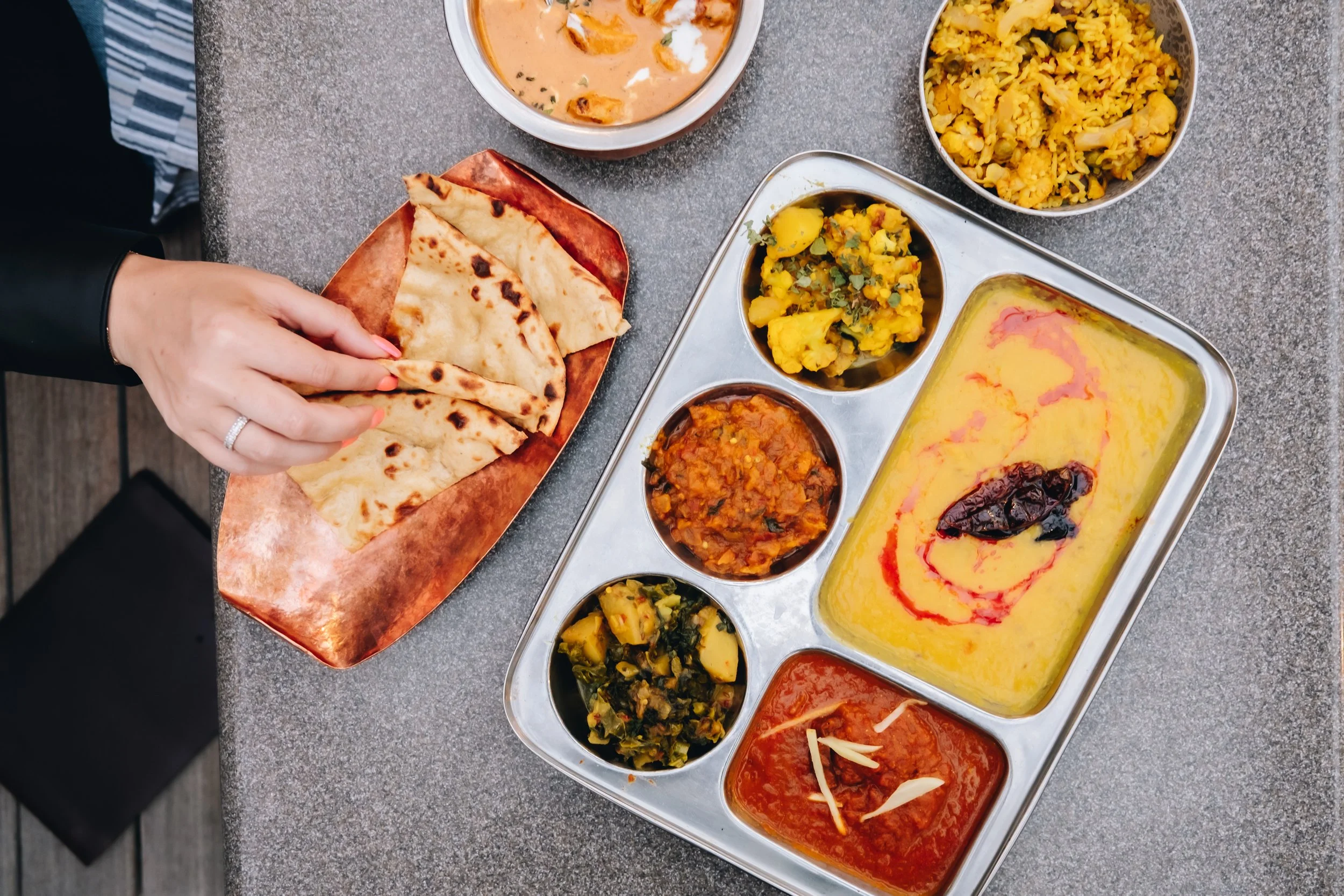 A traditional Indian thali meal on a stainless steel tray with various curries, dal, vegetables, and rice, served with naan bread on a small copper plate. A hand is reaching for a piece of naan. The meal is on a grey table.