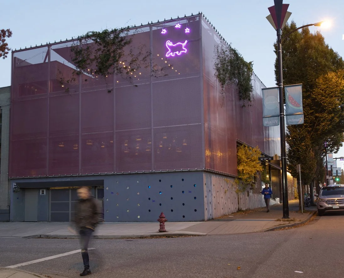A modern building with pink screens and a neon purple dog sign on top, situated on a city street corner during dusk.