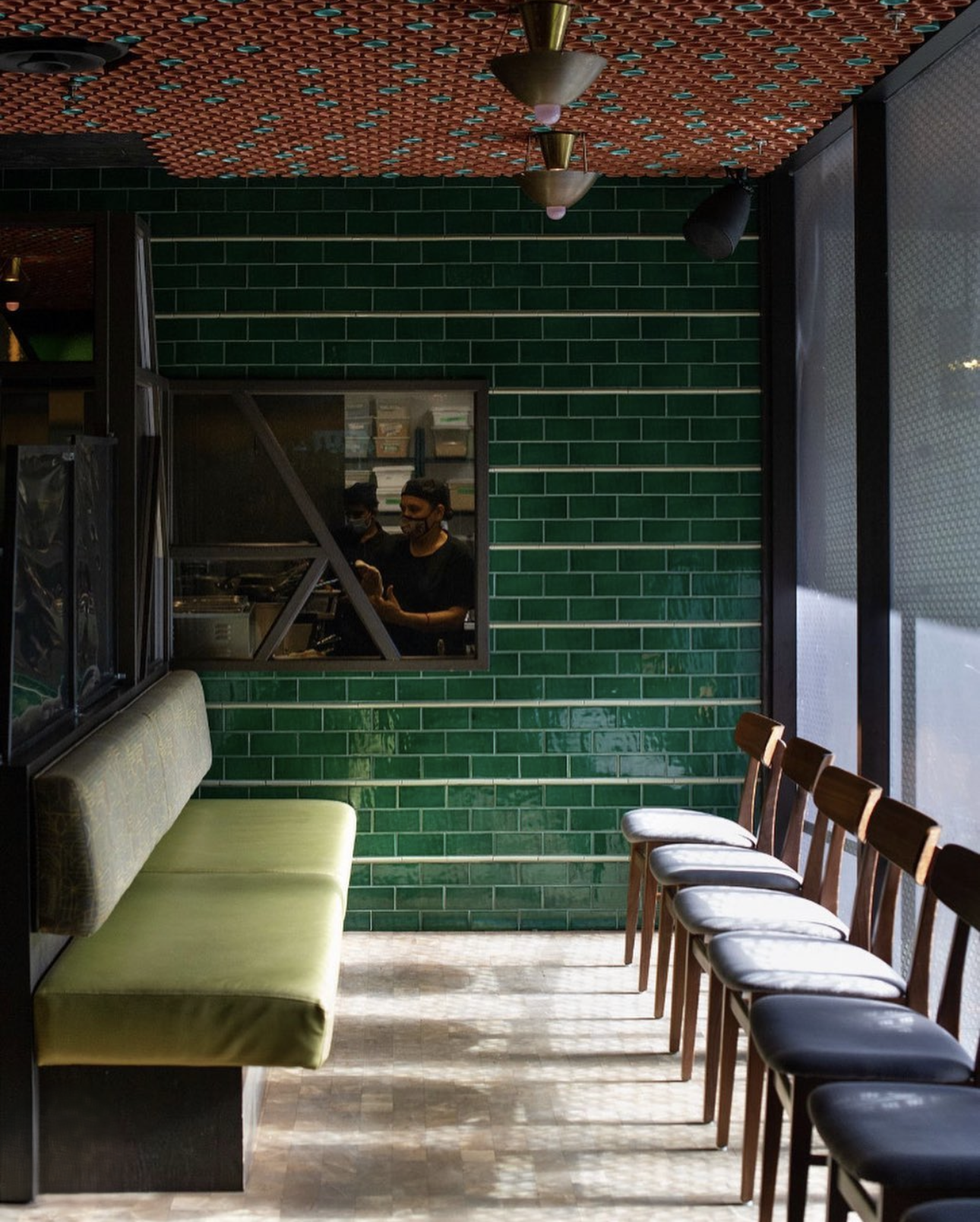 Interior of a restaurant or café with green tiled wall, a row of wooden chairs with black and beige seats, a green cushioned bench, and a window revealing two staff members wearing black masks working behind the counter.