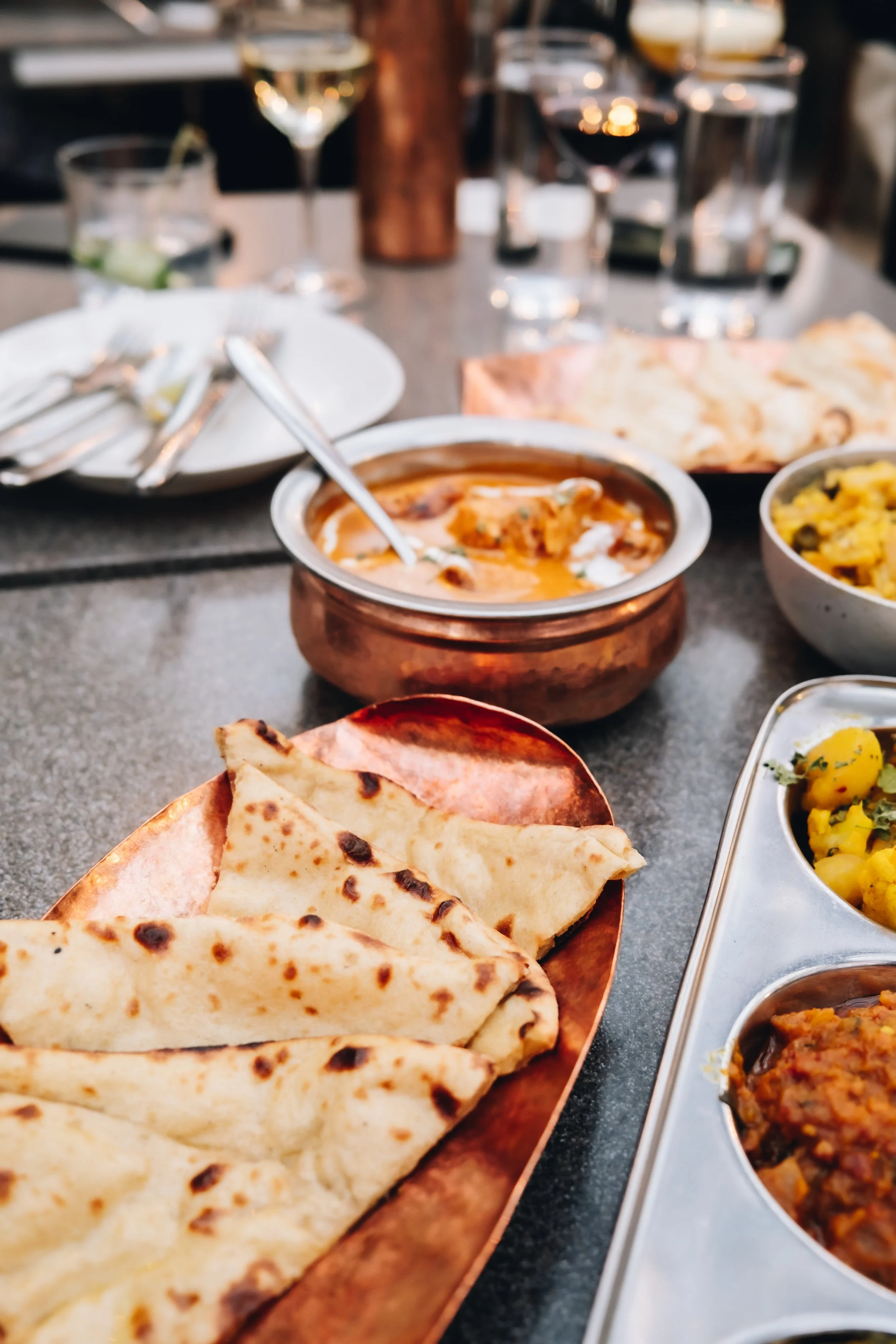 Indian meal with naan bread, creamy curry, vegetable dishes, and drinks on a restaurant table.