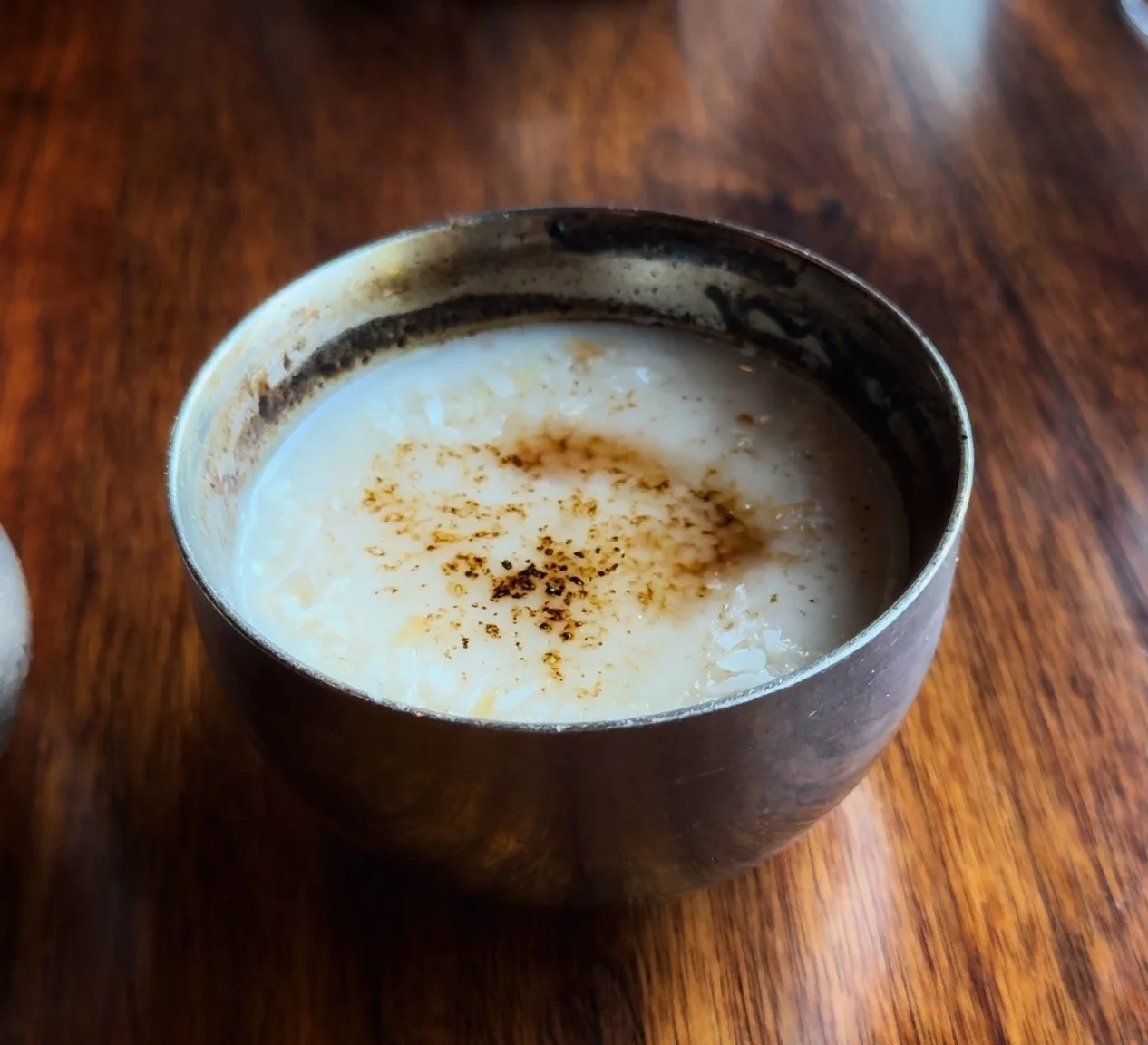 A close-up of a metal cup filled with a creamy drink, topped with cinnamon, sitting on a wooden surface.