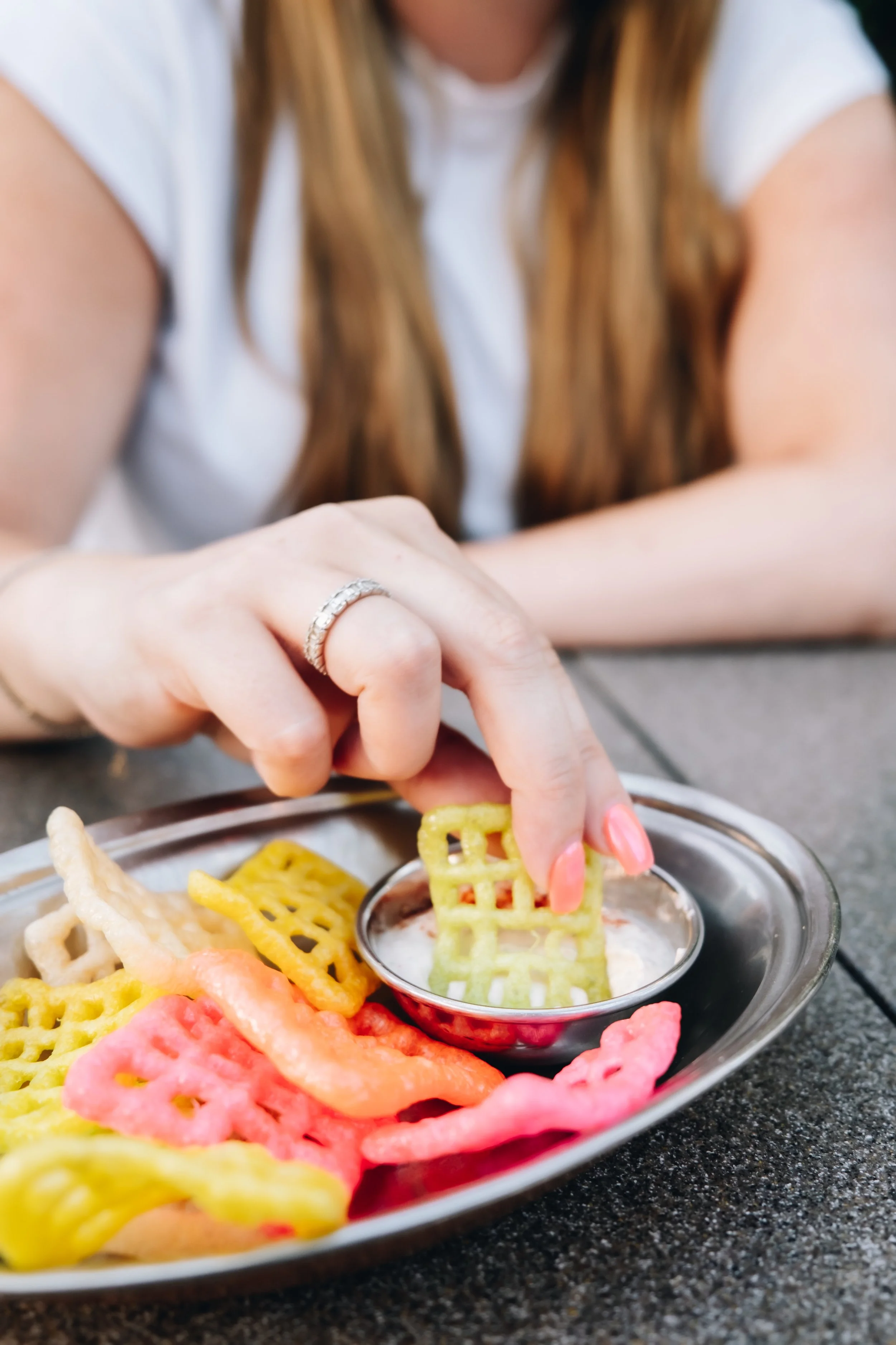 A woman with long brown hair wearing a white shirt dipping colorful waffle chips into a bowl of white dip on a gray table.
