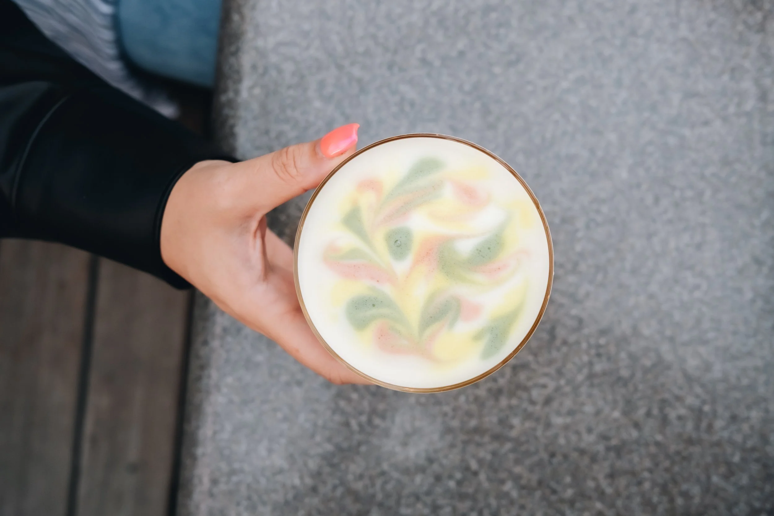 A hand holding a cup of latte with colorful latte art on top, featuring a leaf and swirl pattern in pink, green, and yellow colors.