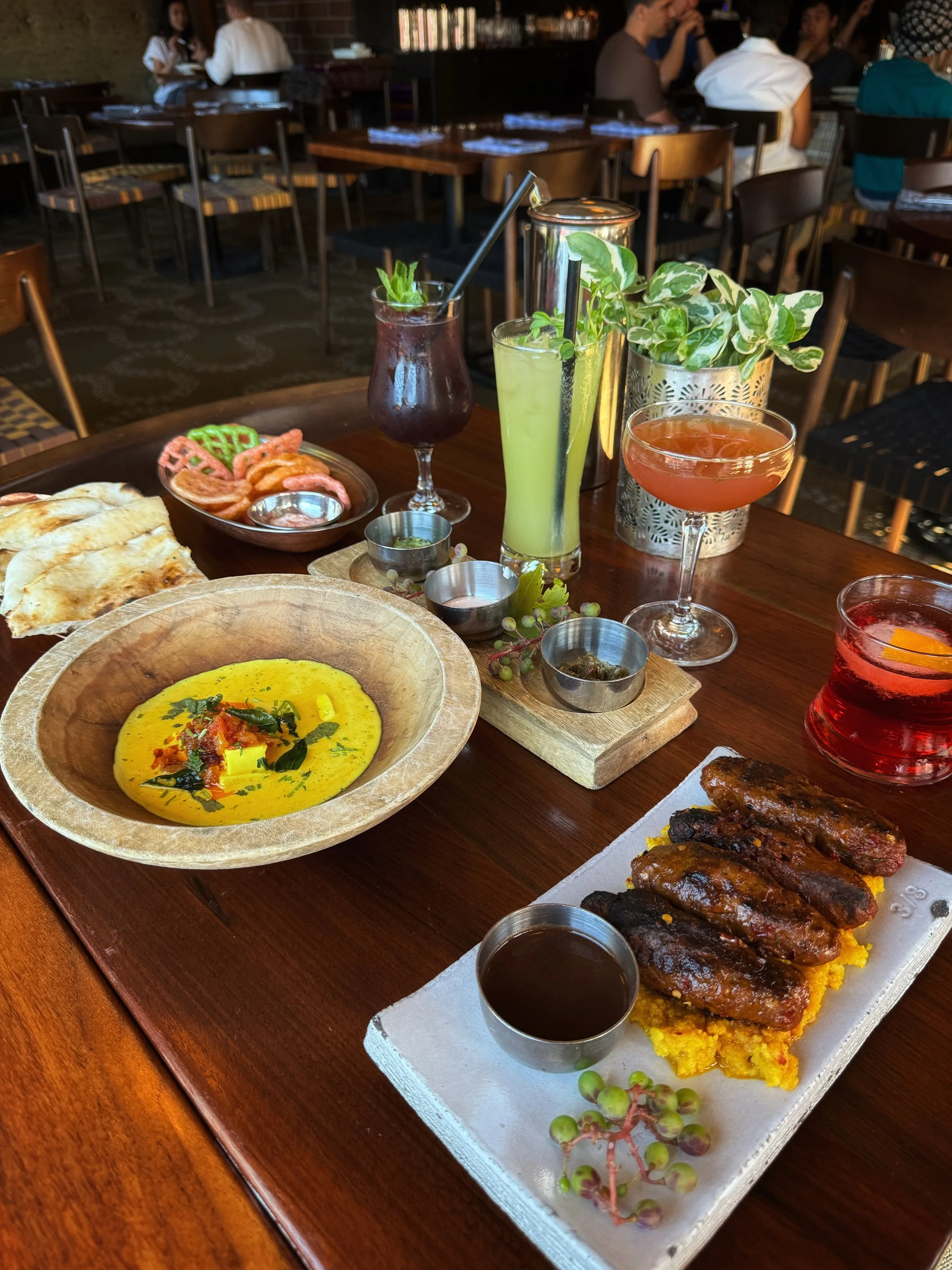 A table at a restaurant with various dishes and colorful cocktails. There is a bowl of yellow curry or soup, a plate with grilled sausages or kebabs over yellow rice, naan bread, and several drinks including a red drink, a green drink, a purple drink