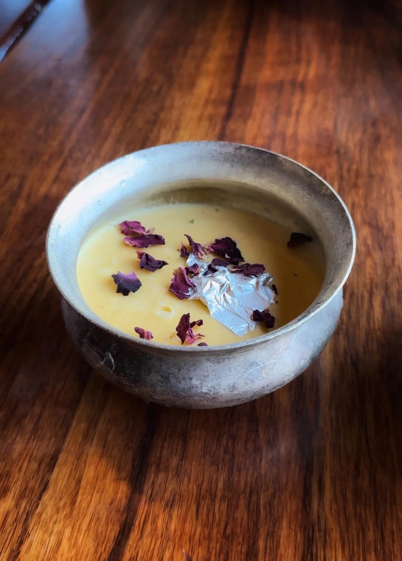 A small bowl of yellow dessert, topped with dried purple flower petals and a piece of crumpled silver foil, placed on a wooden surface.