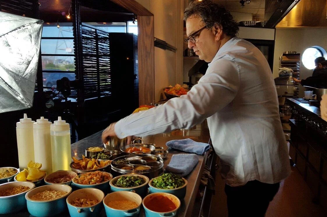 A chef preparing a variety of sauces and dishes in a kitchen. The counter is lined with bowls of different sauces, condiments, and vegetables. The chef is wearing glasses and a white coat, focused on his work. There are kitchen towels and bottles on 