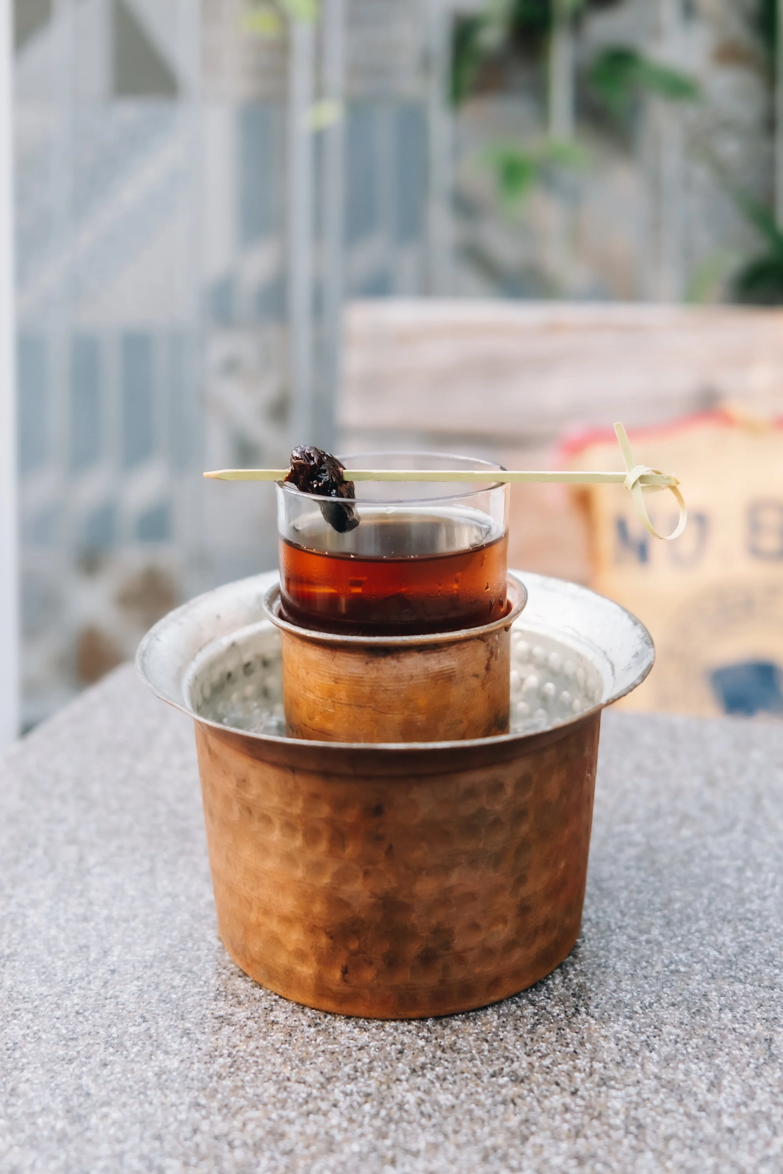A glass of dark beverage on top of a small copper container with hot water, garnished with a piece of dark fruit on a stick.