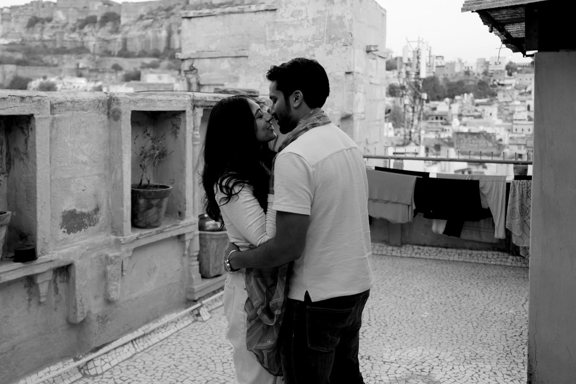 Black and white photo of a couple embracing on a rooftop, with laundry hanging in the background and a cityscape view.