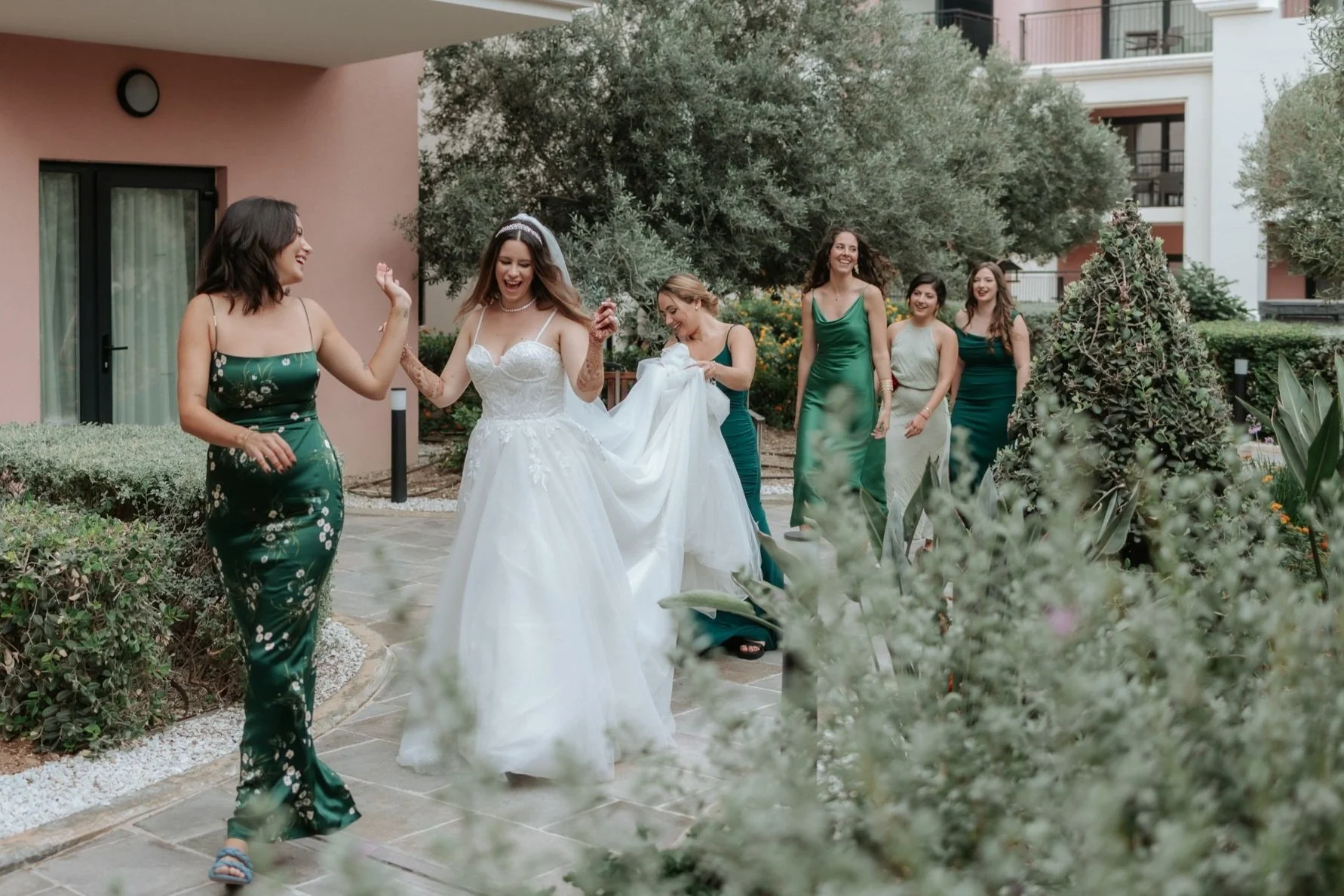 Bride and bridesmaids walking outdoors, bride in white gown, bridesmaids in green dresses, joyful and smiling.