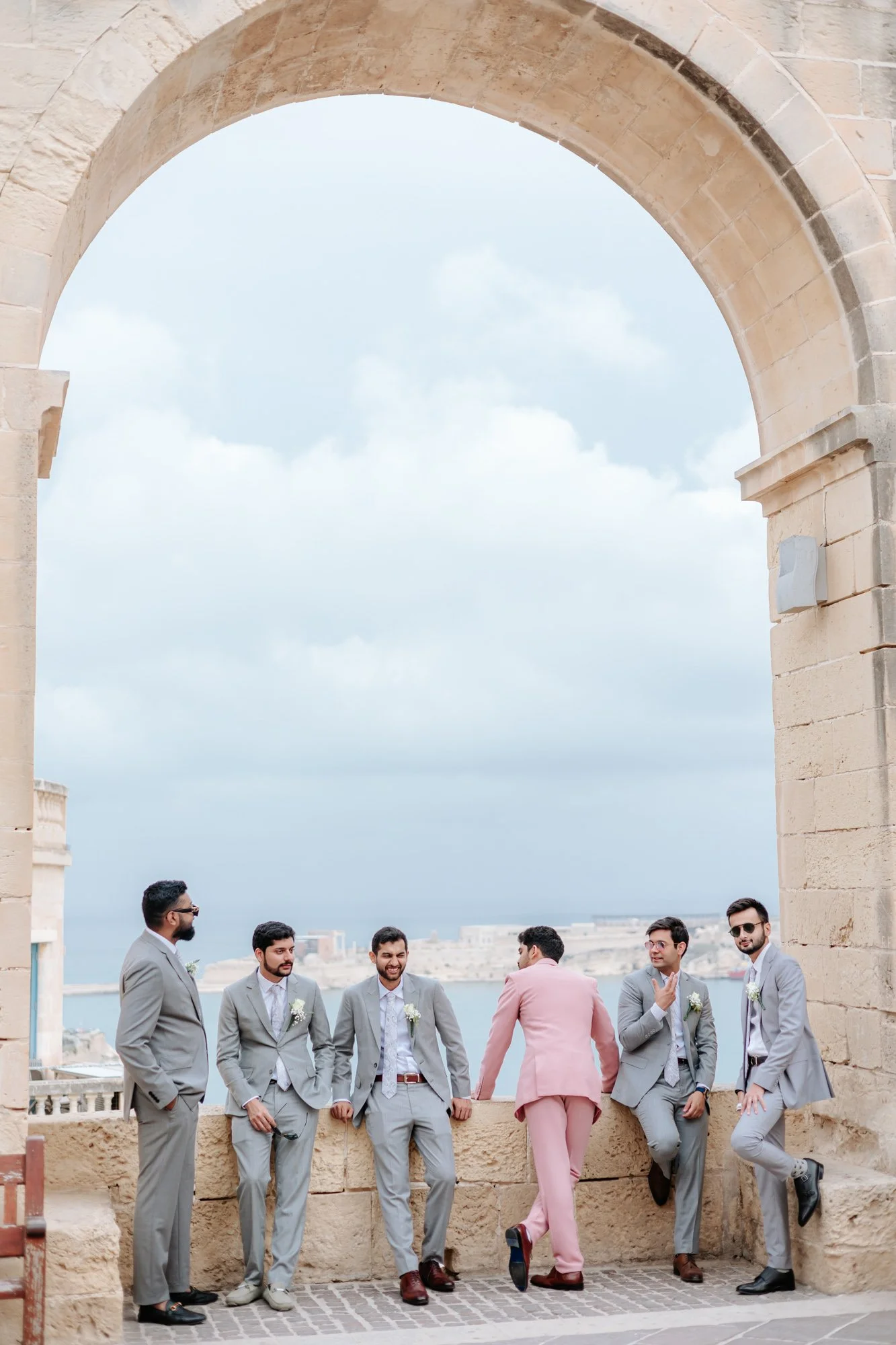 Group of men in suits posing under an archway with a city and ocean view in the background.