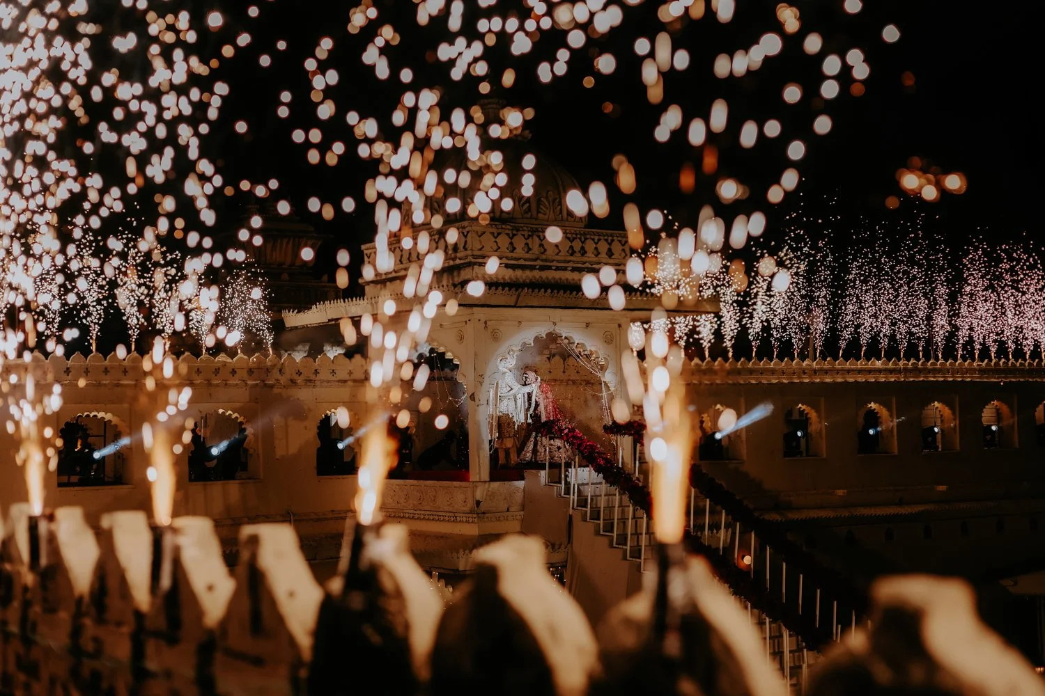 Indian wedding at night with fireworks, traditional architecture, and a bride and groom.