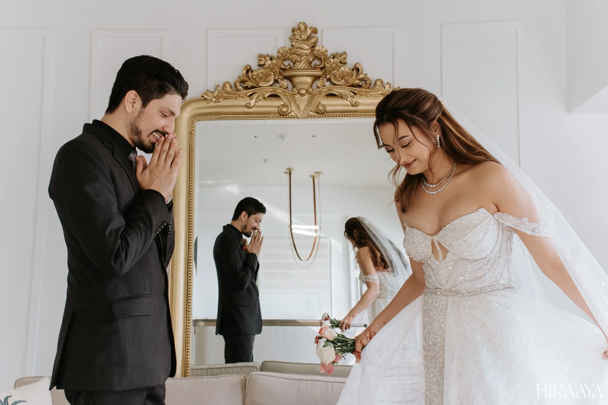 Bride and groom preparing for wedding, the bride in a white dress holding a bouquet and the groom in a black suit, standing near a decorative mirror showing their reflection.