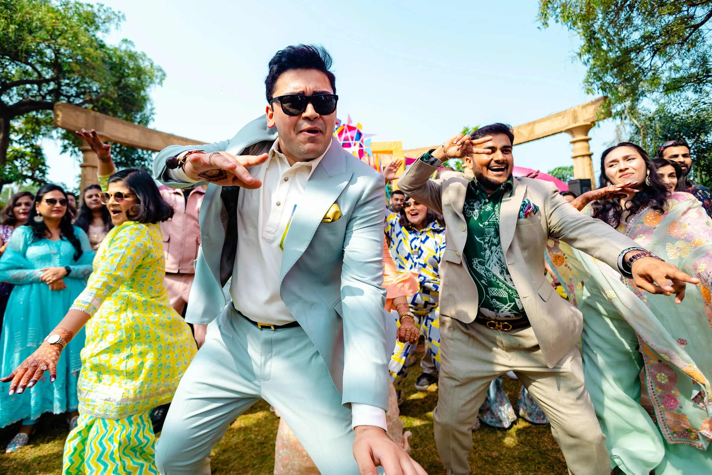 People dancing energetically at an outdoor celebration, dressed in colorful and formal attire, under a clear sky.