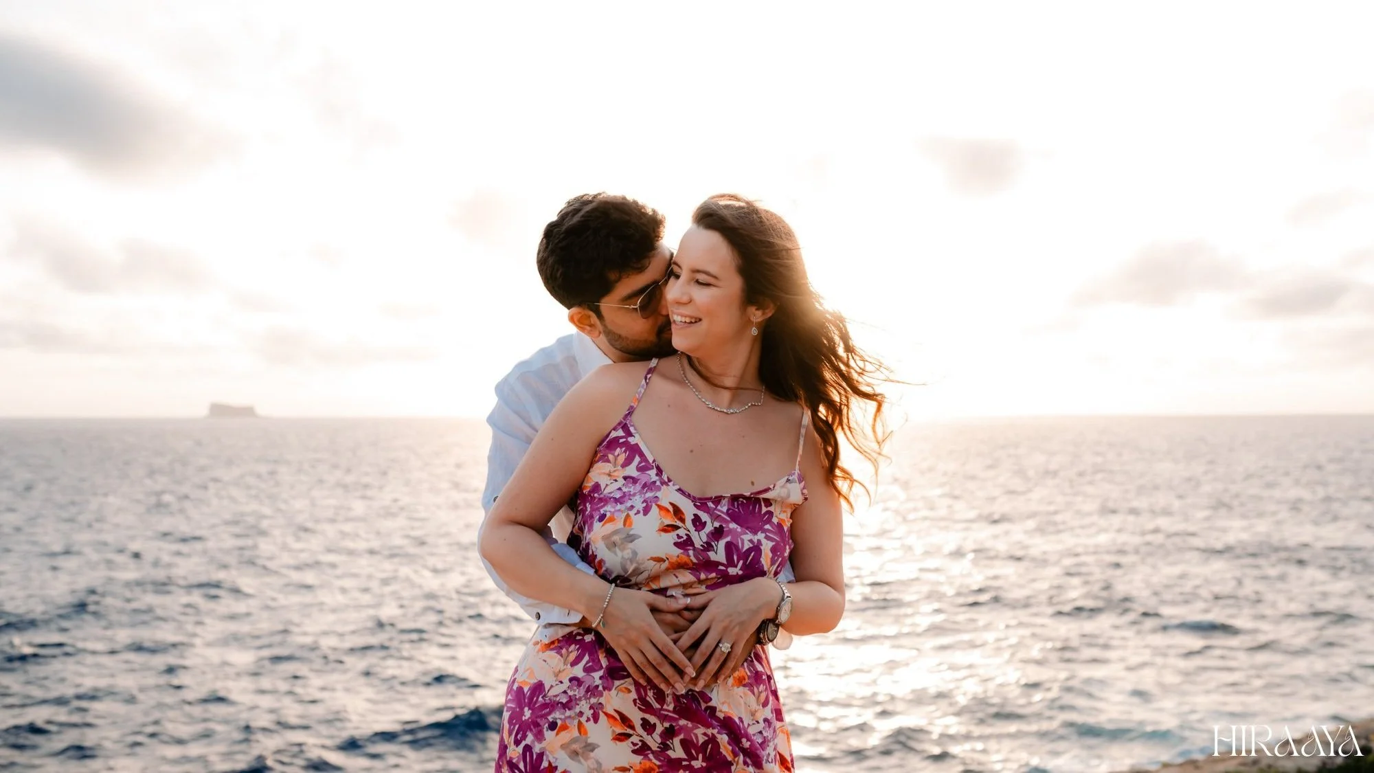 A couple embracing by the ocean during sunset, woman wearing a floral dress, man in a white shirt.