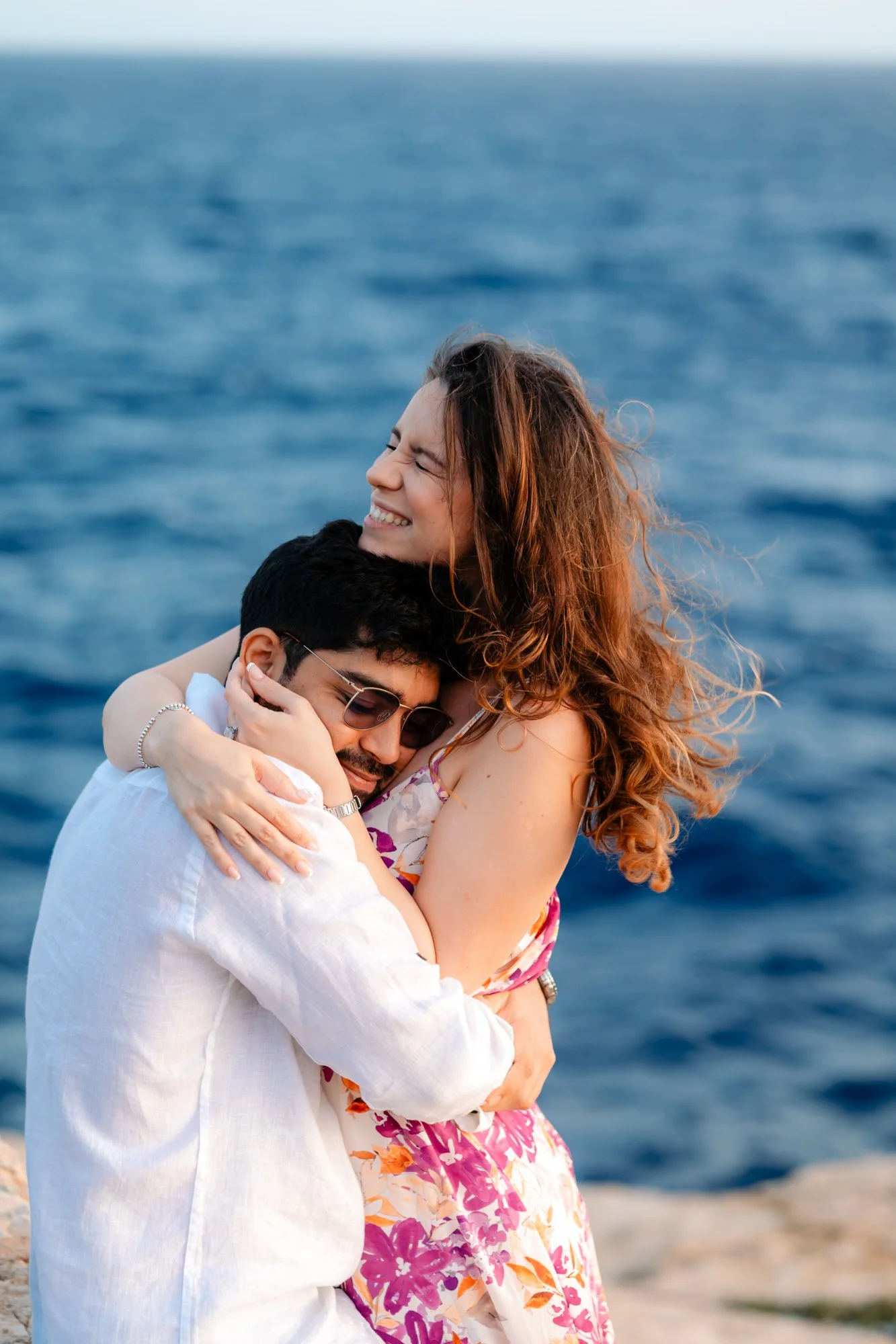 A couple embracing near the ocean, with the woman smiling and wearing a floral dress. The man is in a white shirt and sunglasses.