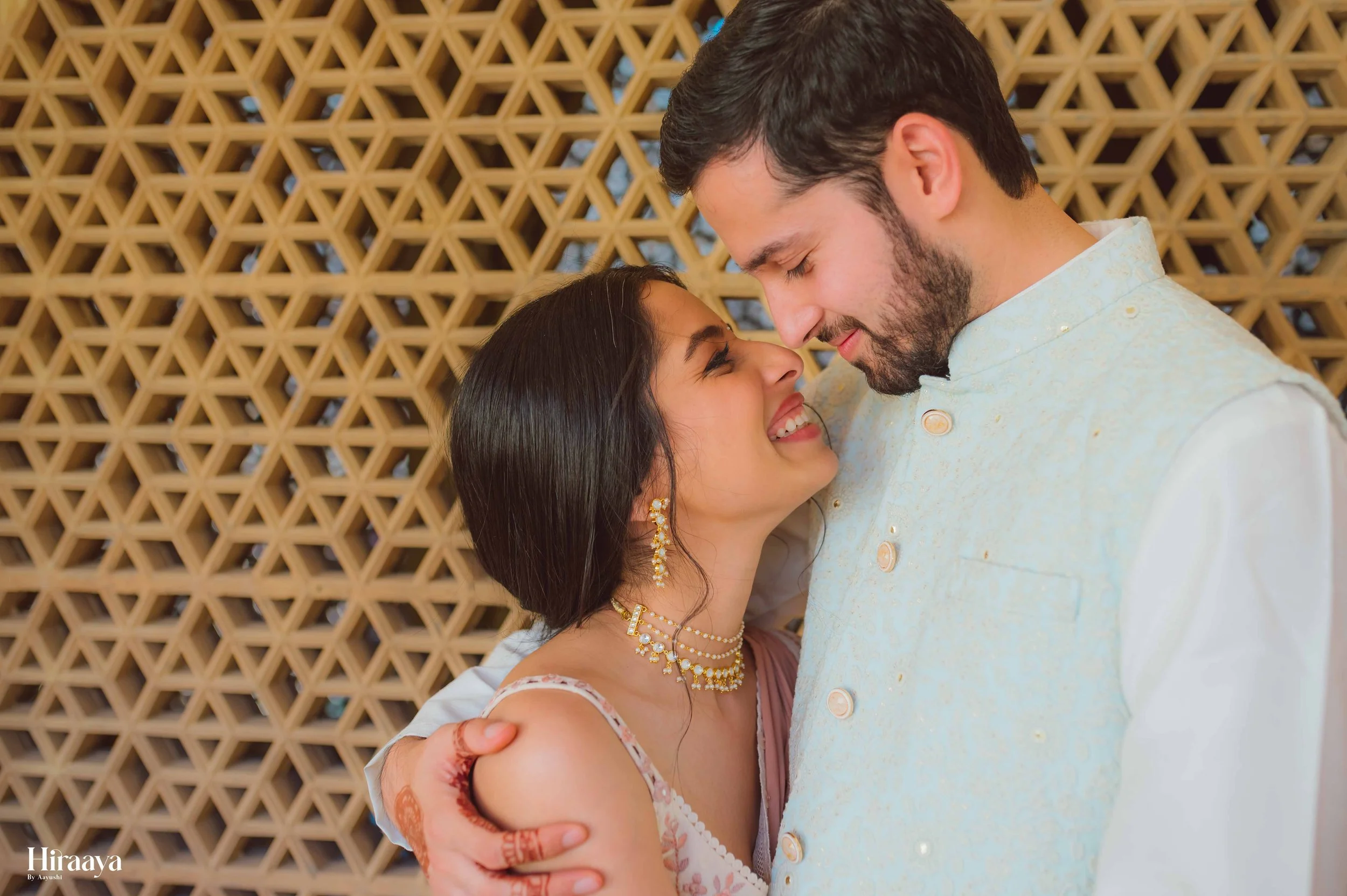 A smiling couple in traditional attire embracing each other with their foreheads touching; the woman is wearing a multistrand necklace and earrings, with a henna design on her hand. The background features a geometric pattern.