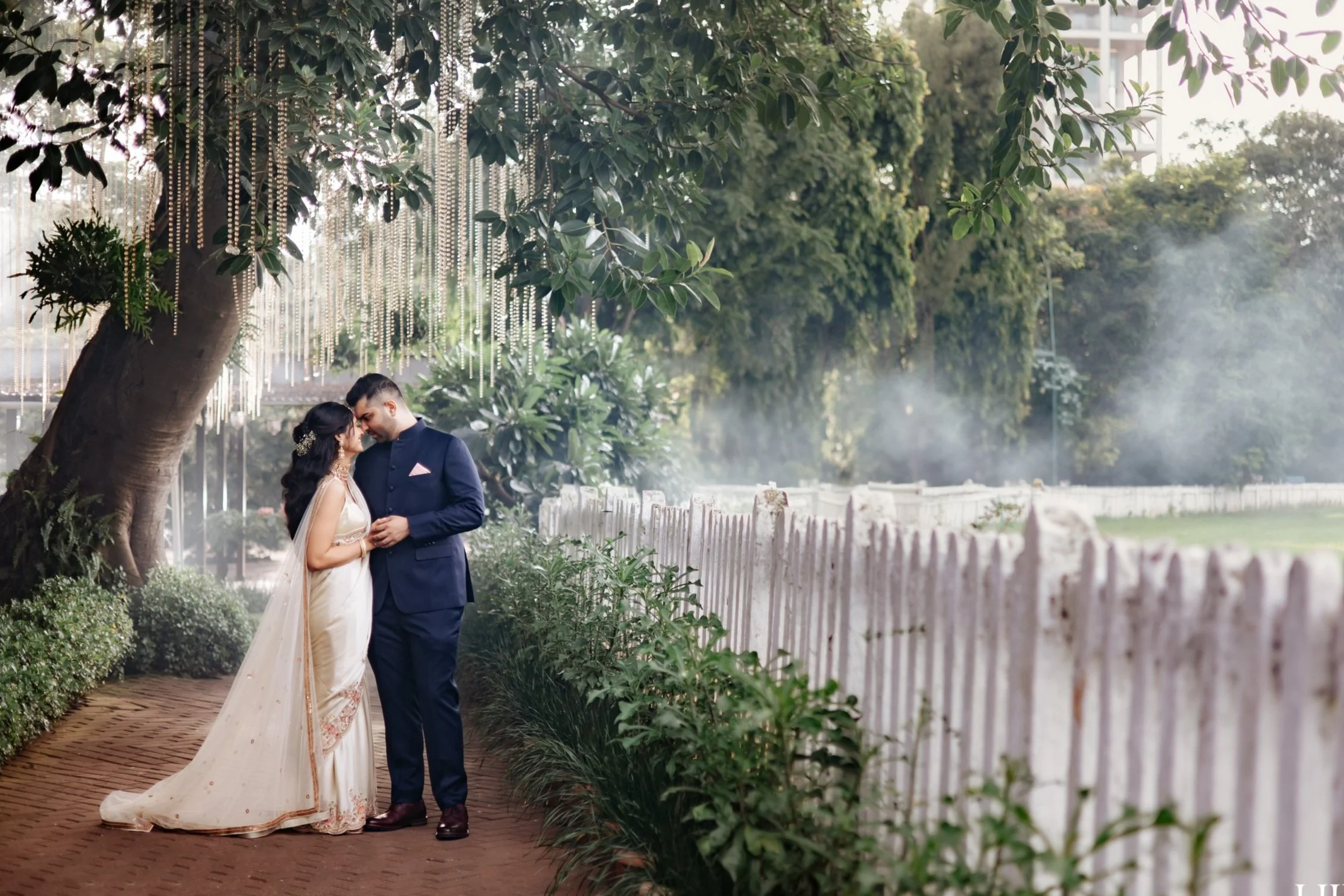 A couple stands close together in a garden setting, under a large tree with hanging decorations. The woman wears a traditional saree and the man is in a dark suit. A wooden fence and lush greenery are in the background.