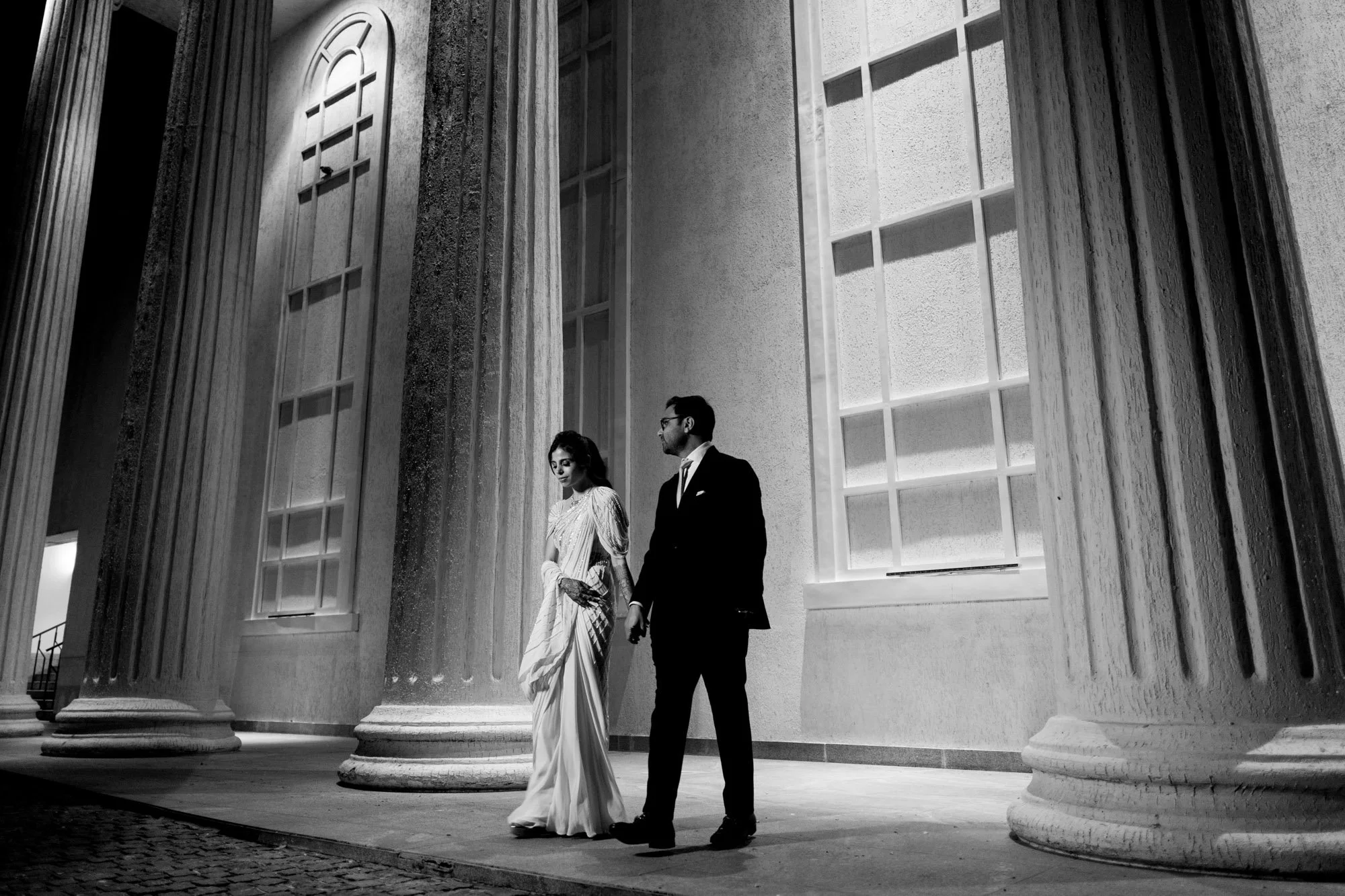 A black and white photo of a couple standing in front of grand architectural columns. The woman is wearing a long dress and the man is in a suit. The setting appears to be a formal, historic building.