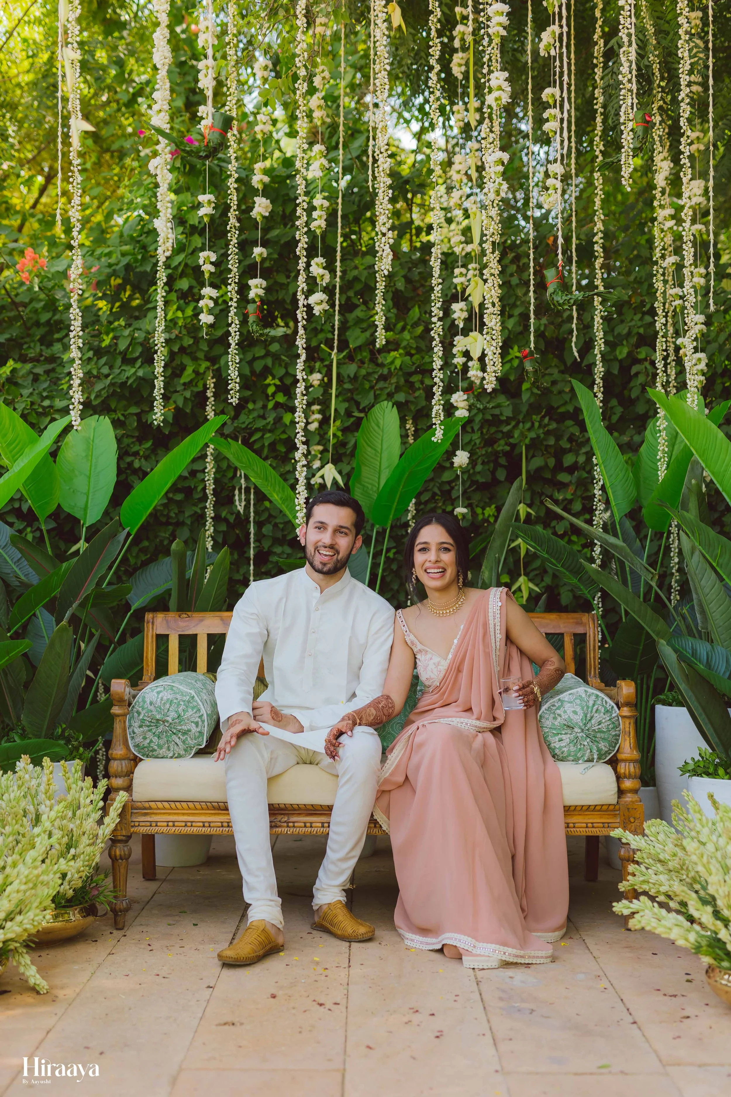 Smiling couple seated on a wooden bench in a garden, dressed in traditional attire with decorative floral strings hanging above, surrounded by lush greenery and potted plants.