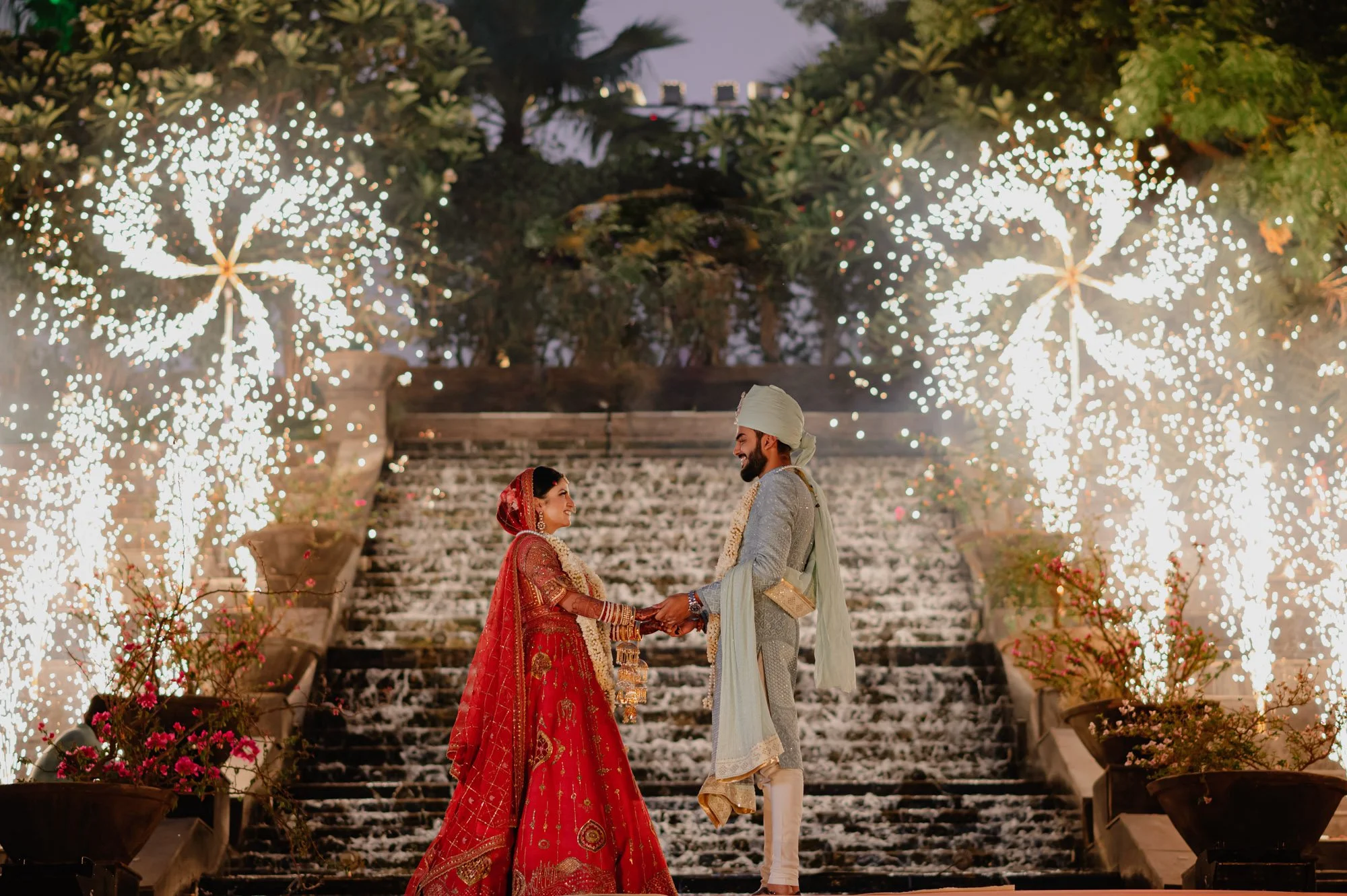 A couple in traditional Indian clothing holding hands in front of a waterfall with fireworks in the background at night.