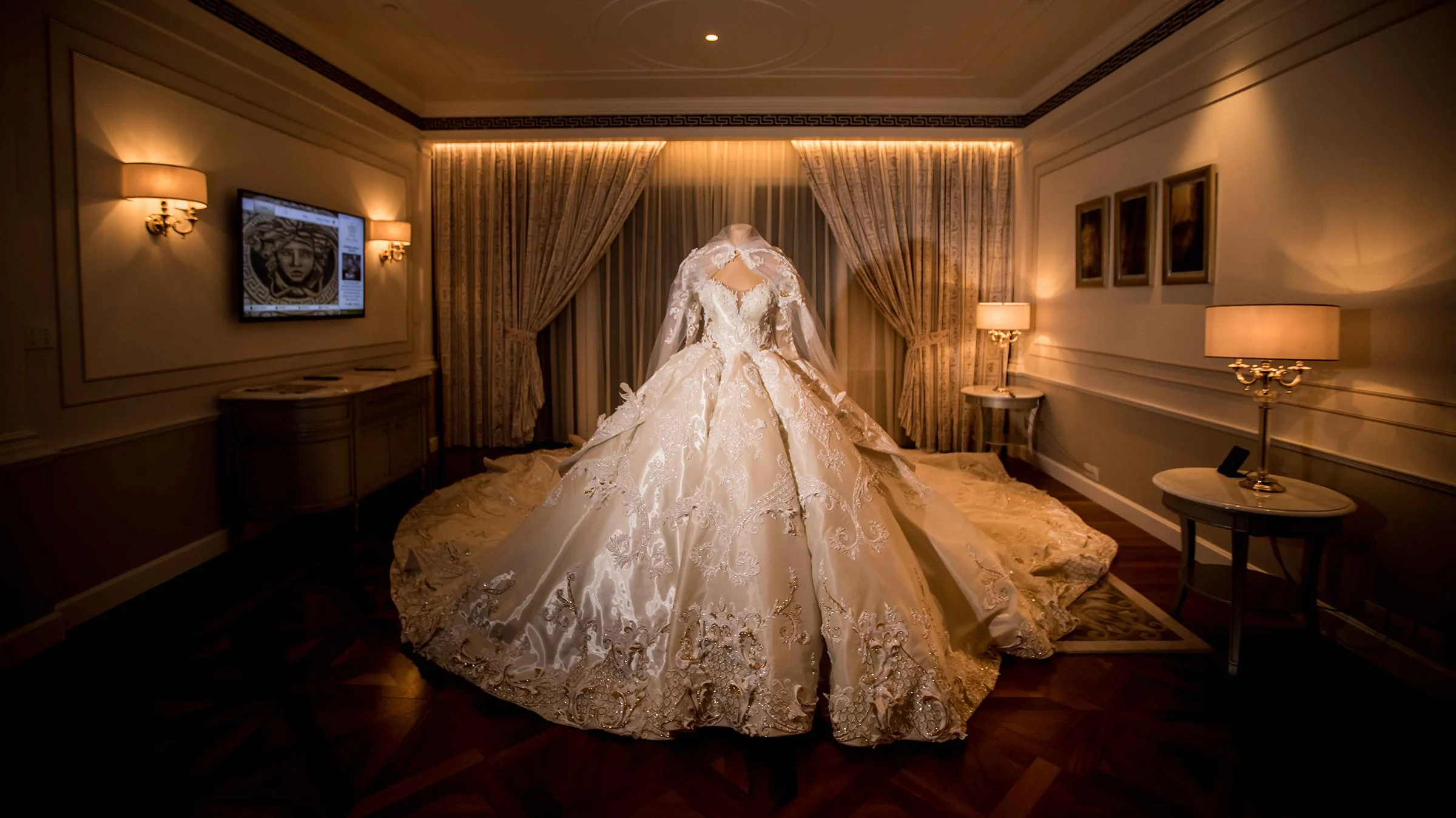 Elegant room with a white, ornate wedding dress displayed on a mannequin, surrounded by soft lighting and luxurious furnishings.
