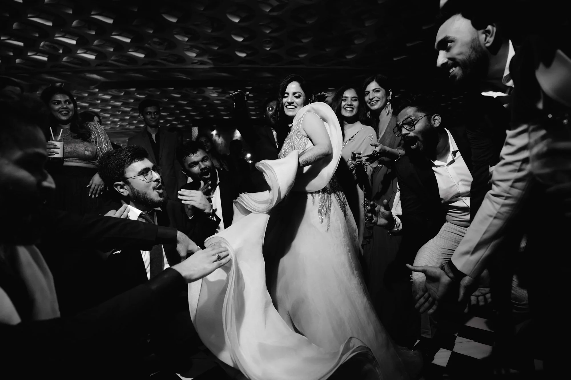 Black and white photo of a group of people celebrating at a party, a woman in a flowing dress dancing in the center, surrounded by smiling and clapping attendees.