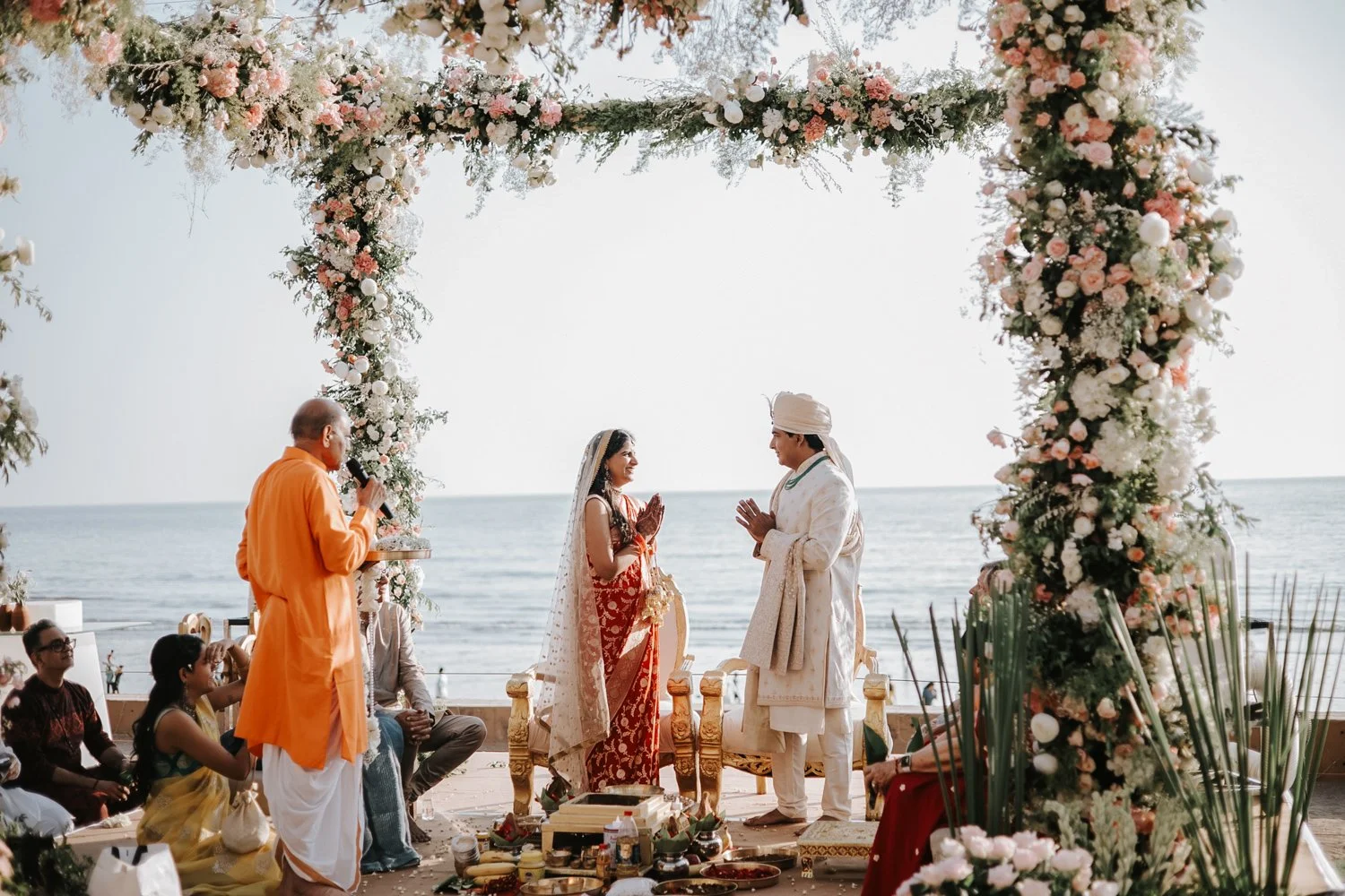 Indian wedding ceremony by the ocean with a couple in traditional attire under a floral arch, surrounded by guests and a priest.