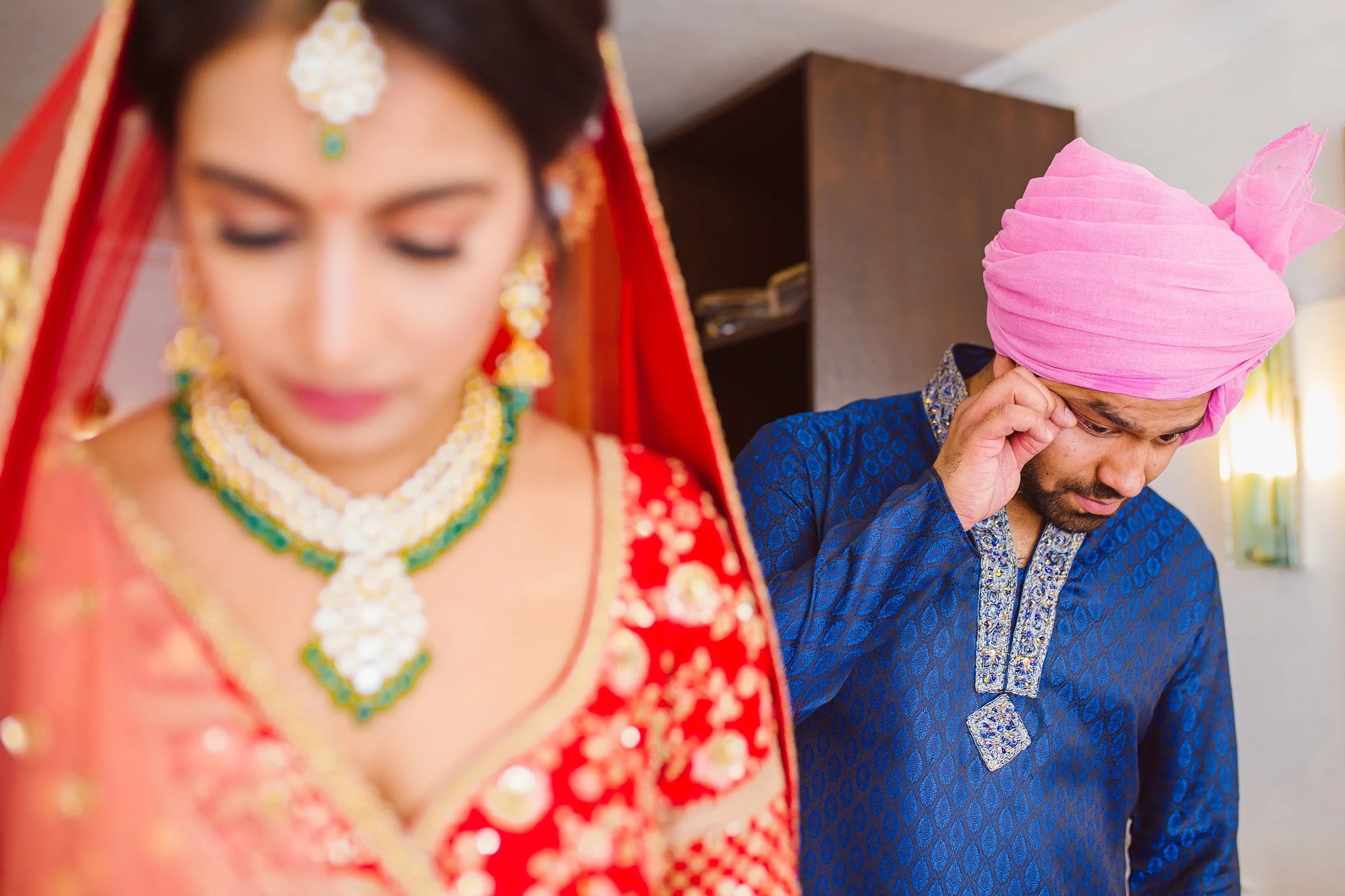 A woman in a red traditional Indian outfit with intricate jewelry is in the foreground, slightly blurred. In the background, a man in a blue outfit with a pink turban is wiping his eye.