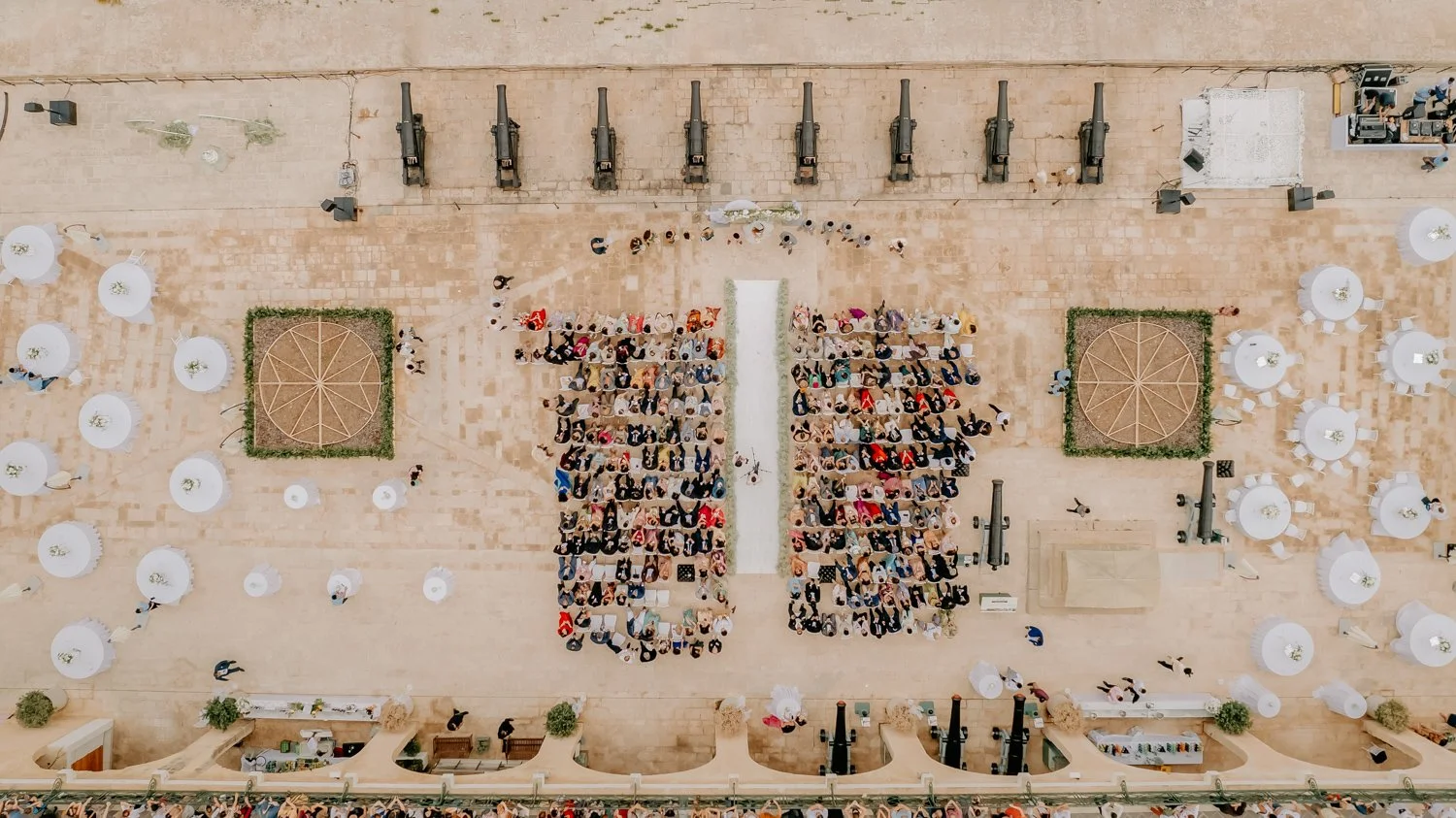 Hiraaya by Aayushi, malta wedding, Indian Wedding Photography, indian wedding, Aerial view of an outdoor event with rows of seated guests, circular tables with white tablecloths, and cannons lined up on a stone courtyard.