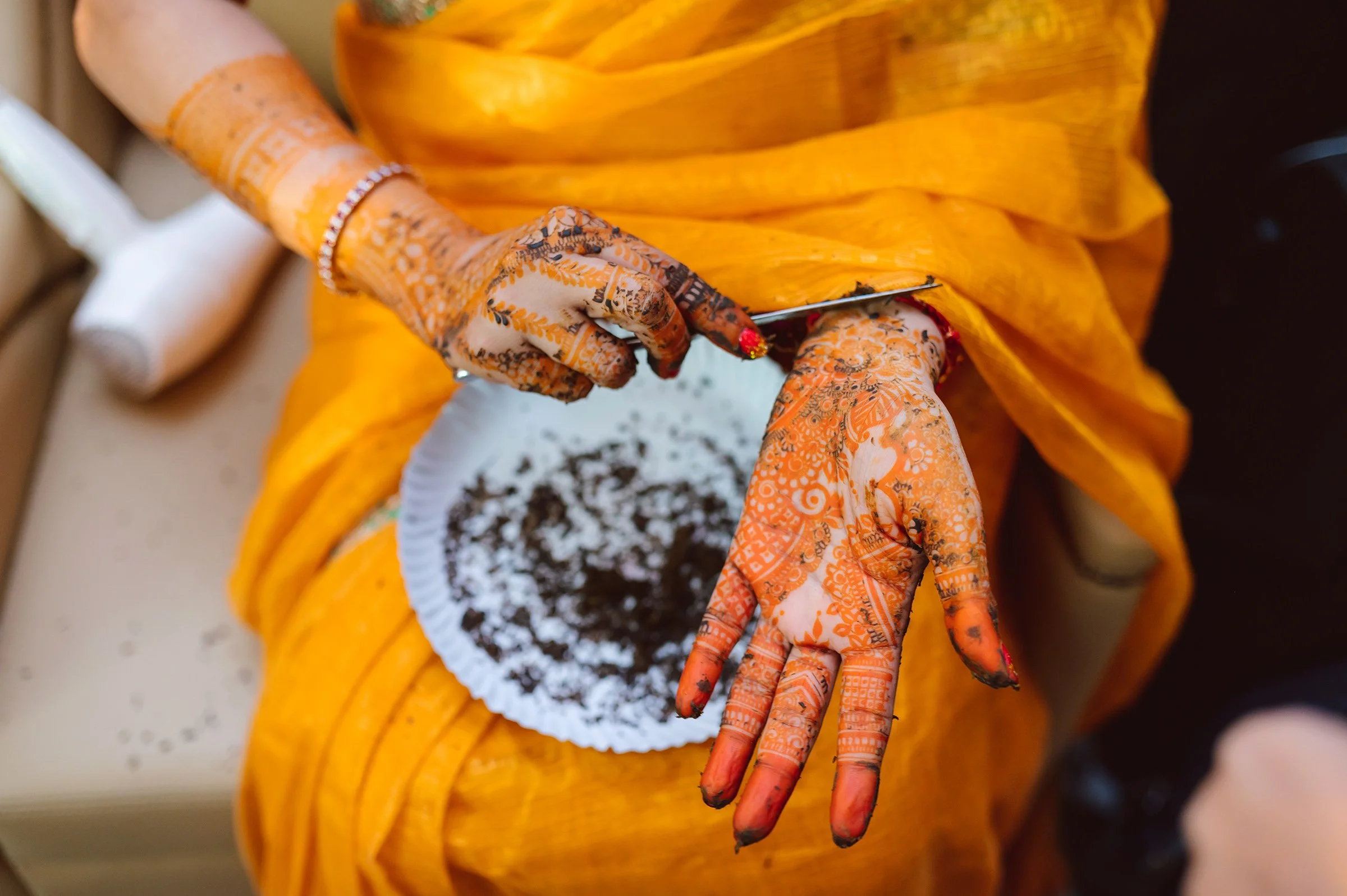 Close-up of a person with henna design on their hands, removing dried henna paste with a knife, wearing an orange sari.