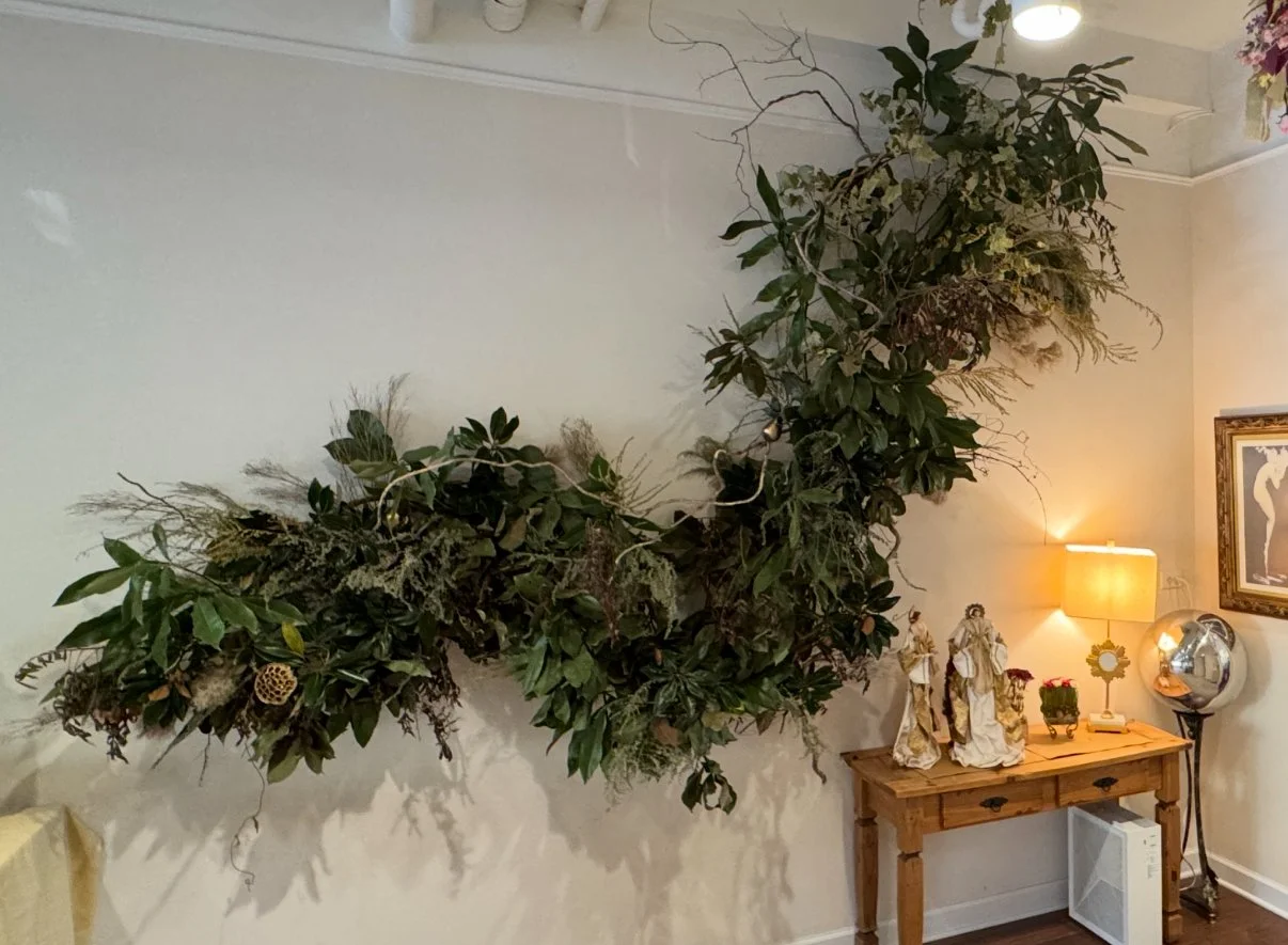 Front steps of a house decorated with pumpkins and colorful chrysanthemums for fall. The pumpkins vary in size and color, and are arranged along the stairs, with two on either side of the top of the stairs. The chrysanthemums are in pots on the left side of the stairs, in pink, purple, and orange shades. A small tree and shrubs are also visible near the door, which has a wrought iron decorative security door.
