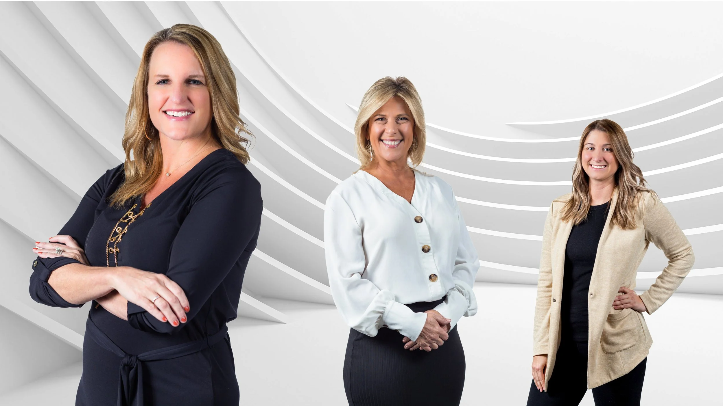 Three women standing in front of a futuristic white background with curved lines. The woman on the left has blonde hair, is wearing a black dress, and has her arms crossed. The woman in the center has blonde hair, is wearing a white blouse with large