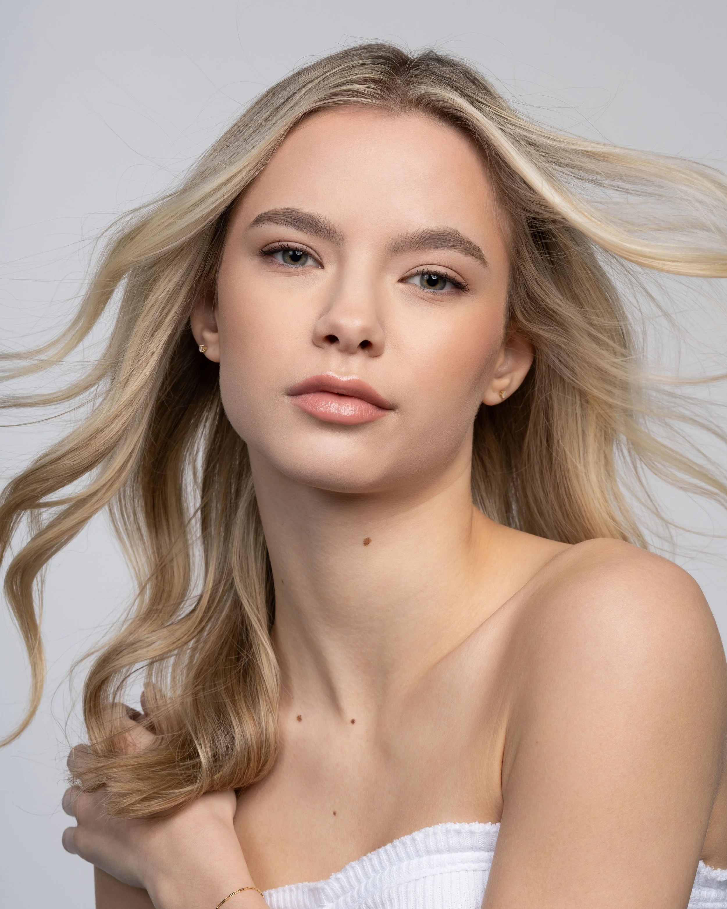 Close-up portrait of a young woman with long, wavy blonde hair, wearing a white strapless top, looking directly at the camera with a neutral expression.