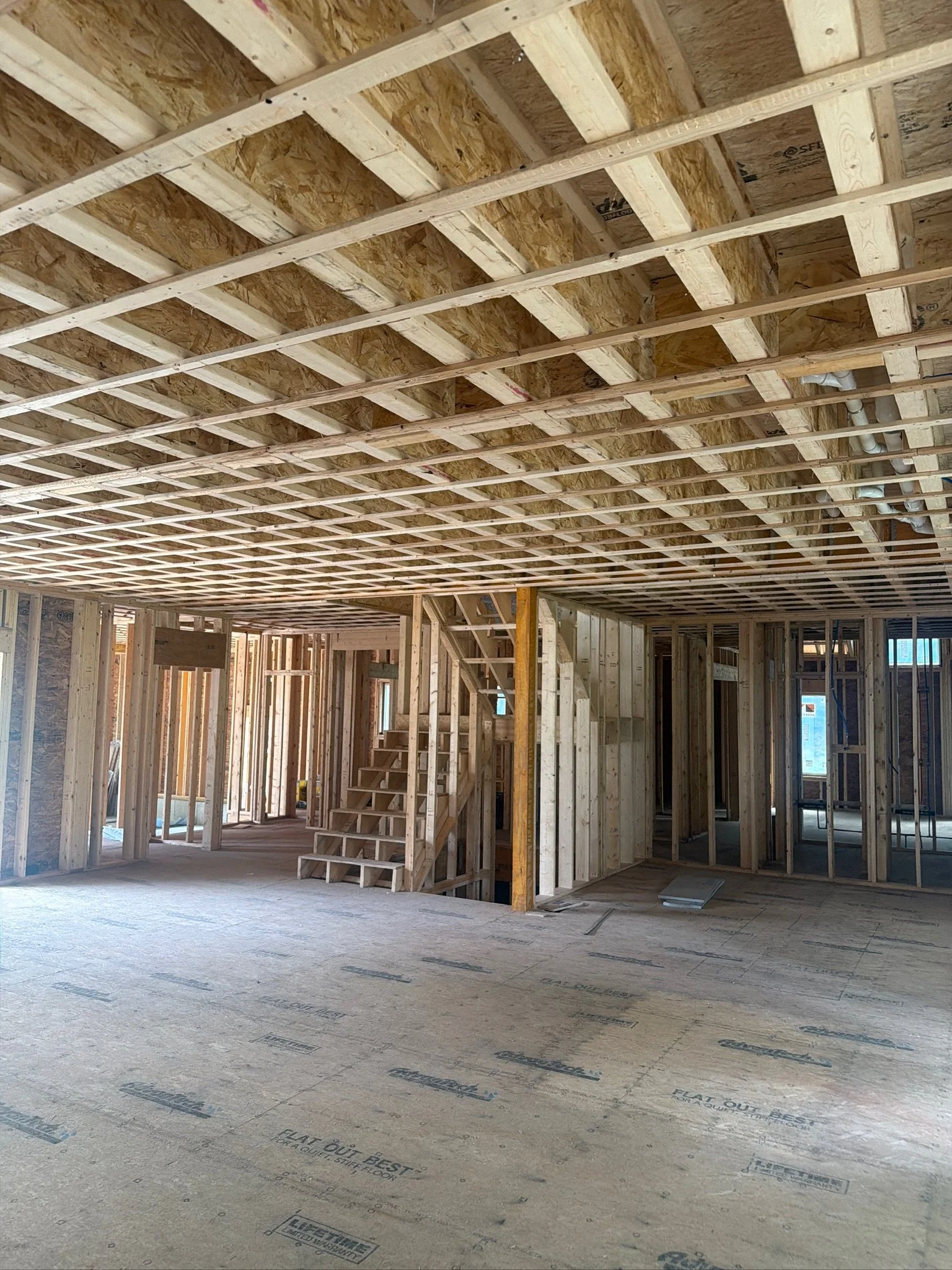 Interior framing progress of a custom home highlighting open sub-flooring, ceiling joists, and wooden stair framing.