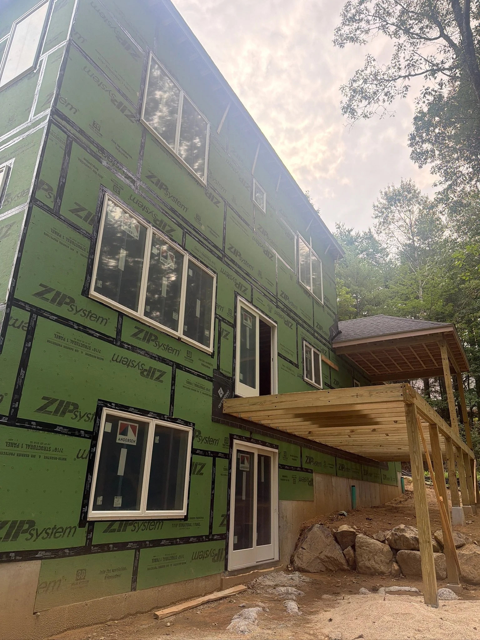 Custom home exterior construction featuring green ZIP System sheathing, installed Andersen windows, and wooden deck support framing.