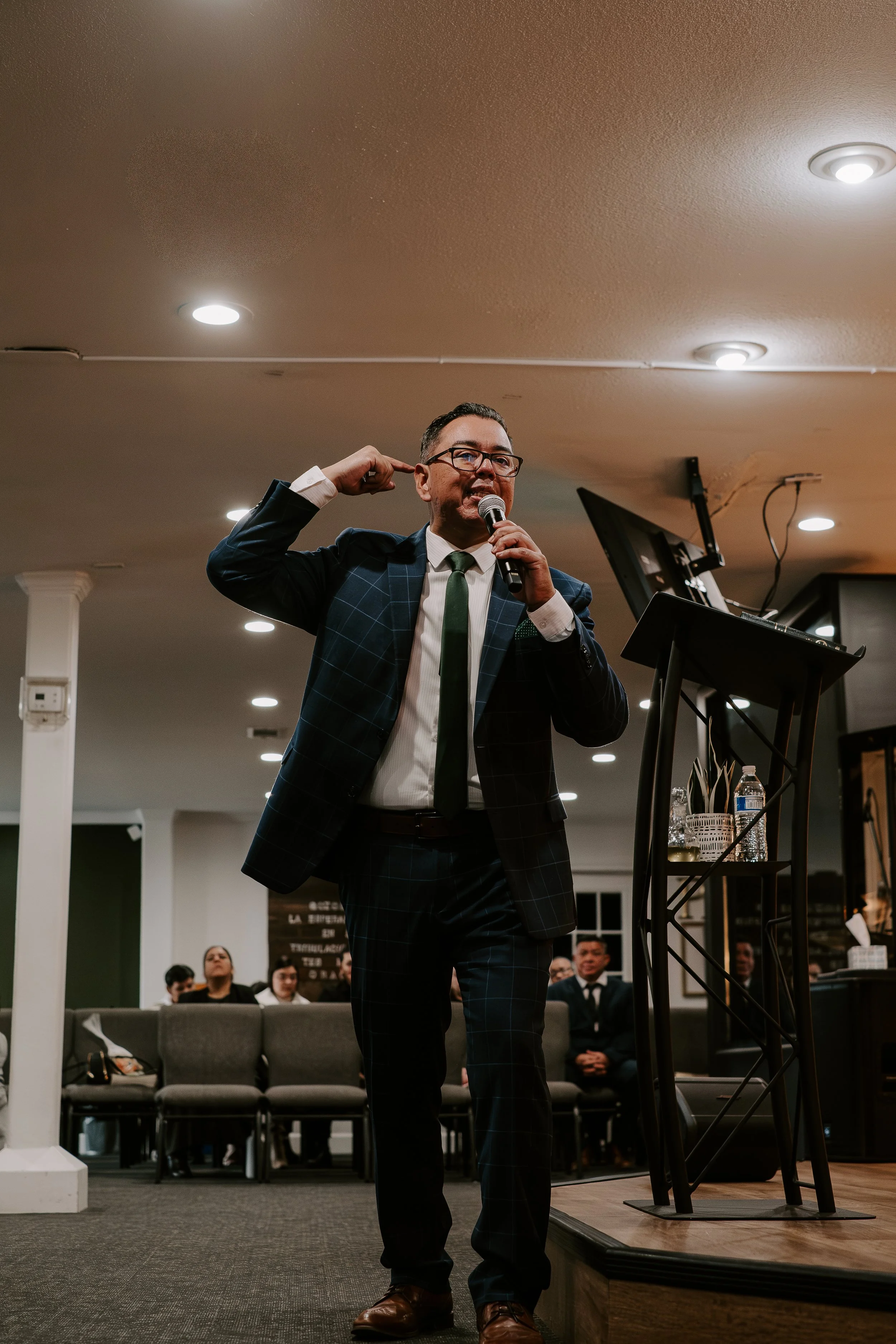A man in a suit and glasses speaking into a microphone at a rally or event, with a microphone stand, water bottle, and audience in the background.