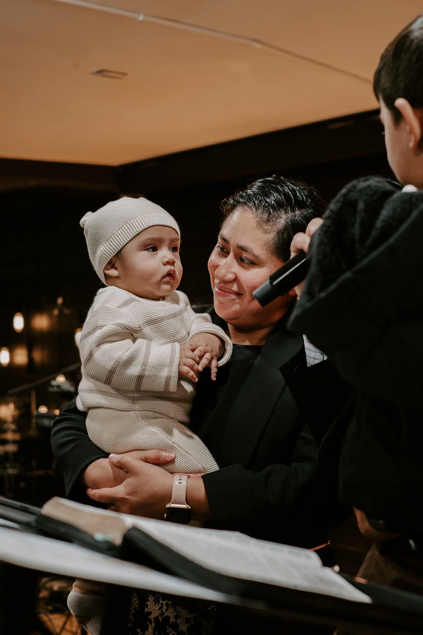 A woman holding a young child and a boy speaking into a microphone in a dimly lit indoor setting.