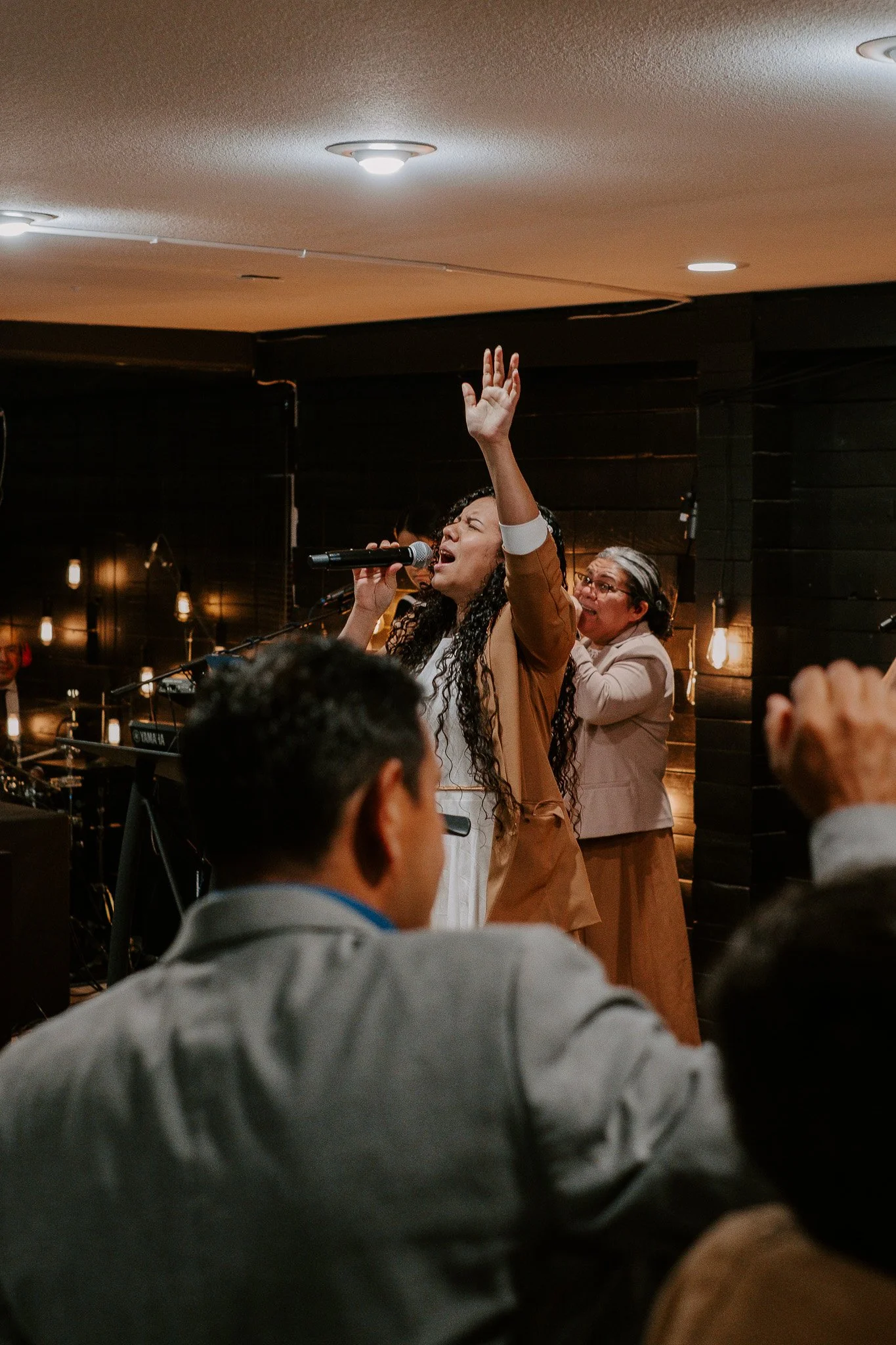 A woman singing passionately into a microphone with her hand raised, surrounded by people in a dimly lit indoor setting.