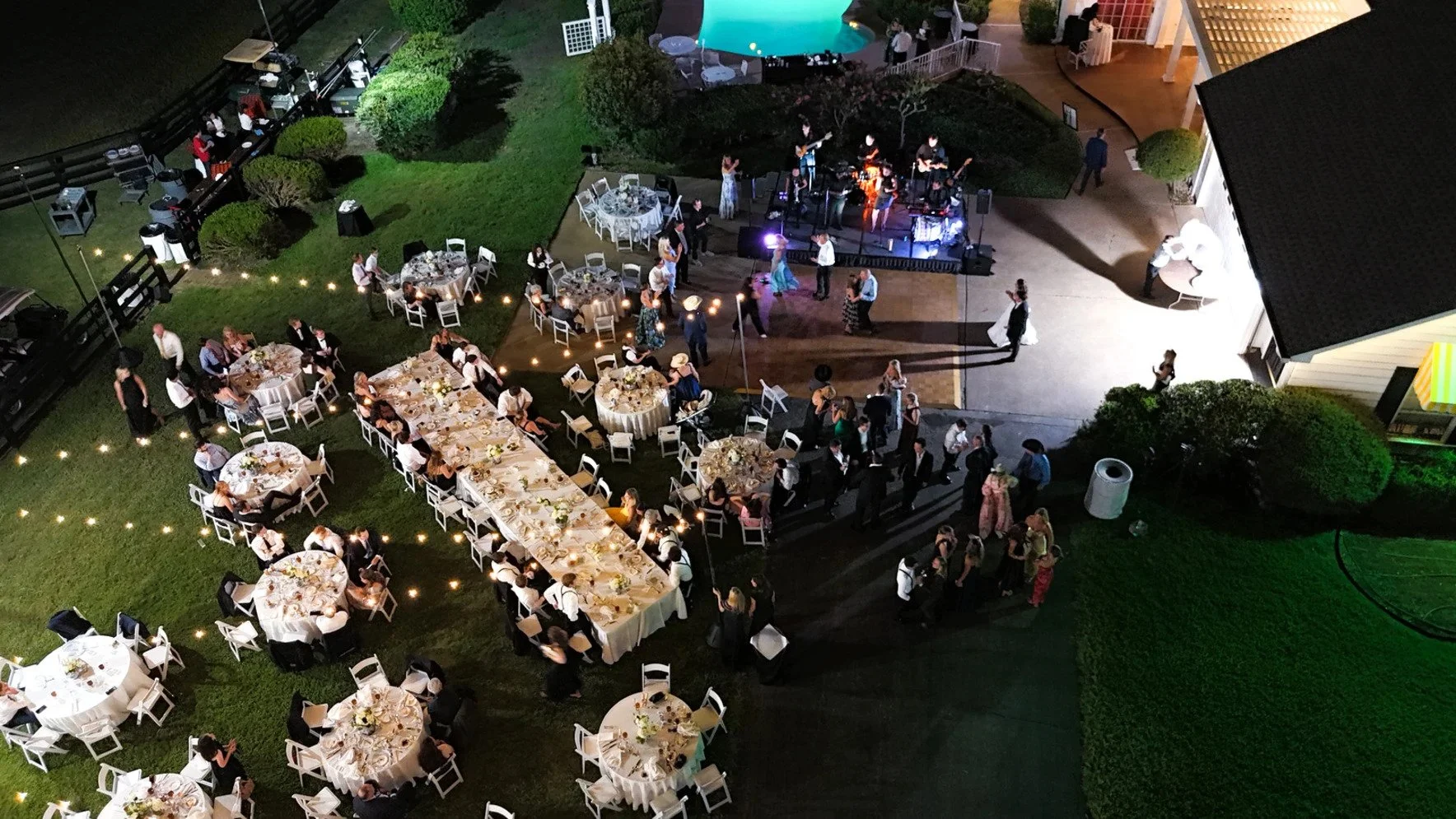 An aerial view of a nighttime outdoor wedding reception with dining tables, a dance floor with live band, and a pool in the background. Guests are seated and dancing, with some standing near the band.