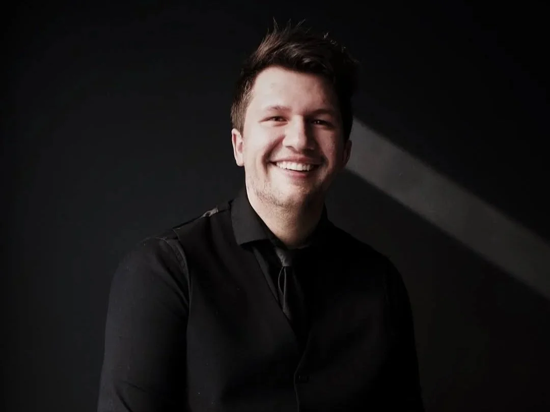 Young man with short brown hair, smiling, wearing a black shirt and black tie against a dark background.