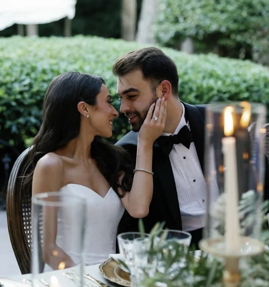 A couple in formal attire sharing an intimate moment at a romantic outdoor dinner, with candles and greenery in the background.