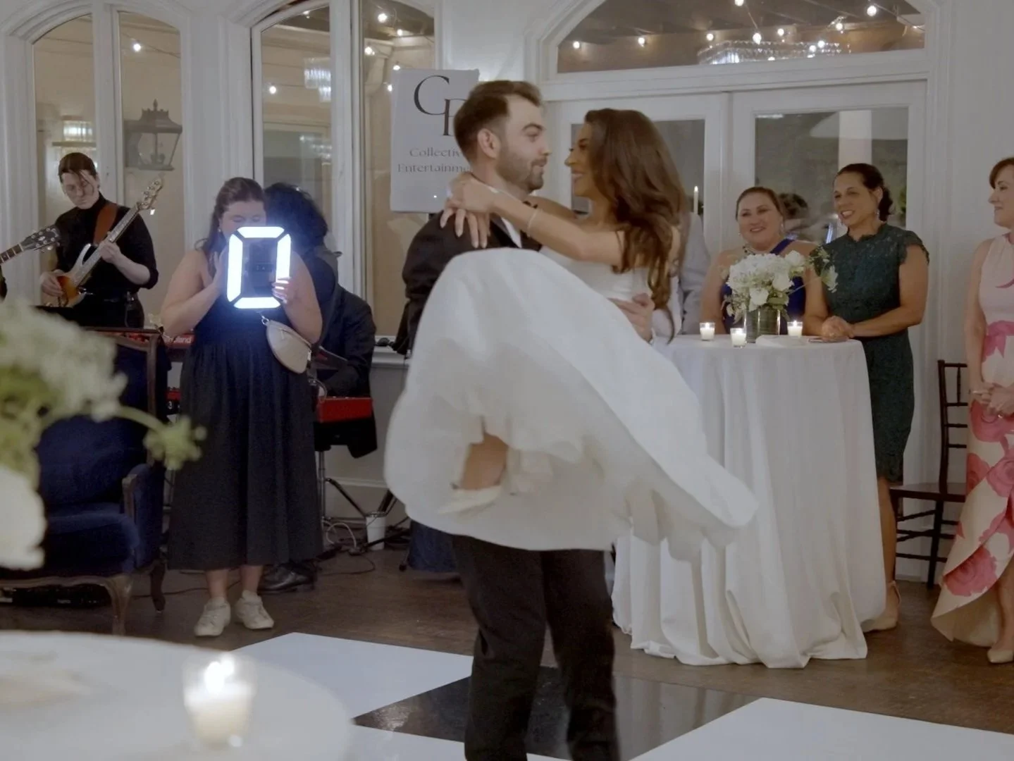 A groom dances with the bride while other guests and a musician watch at a wedding reception in an elegantly decorated room.