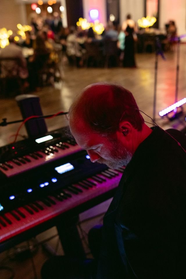 A man playing a keyboard on stage at a lively event or party, with people sitting at tables in the background.