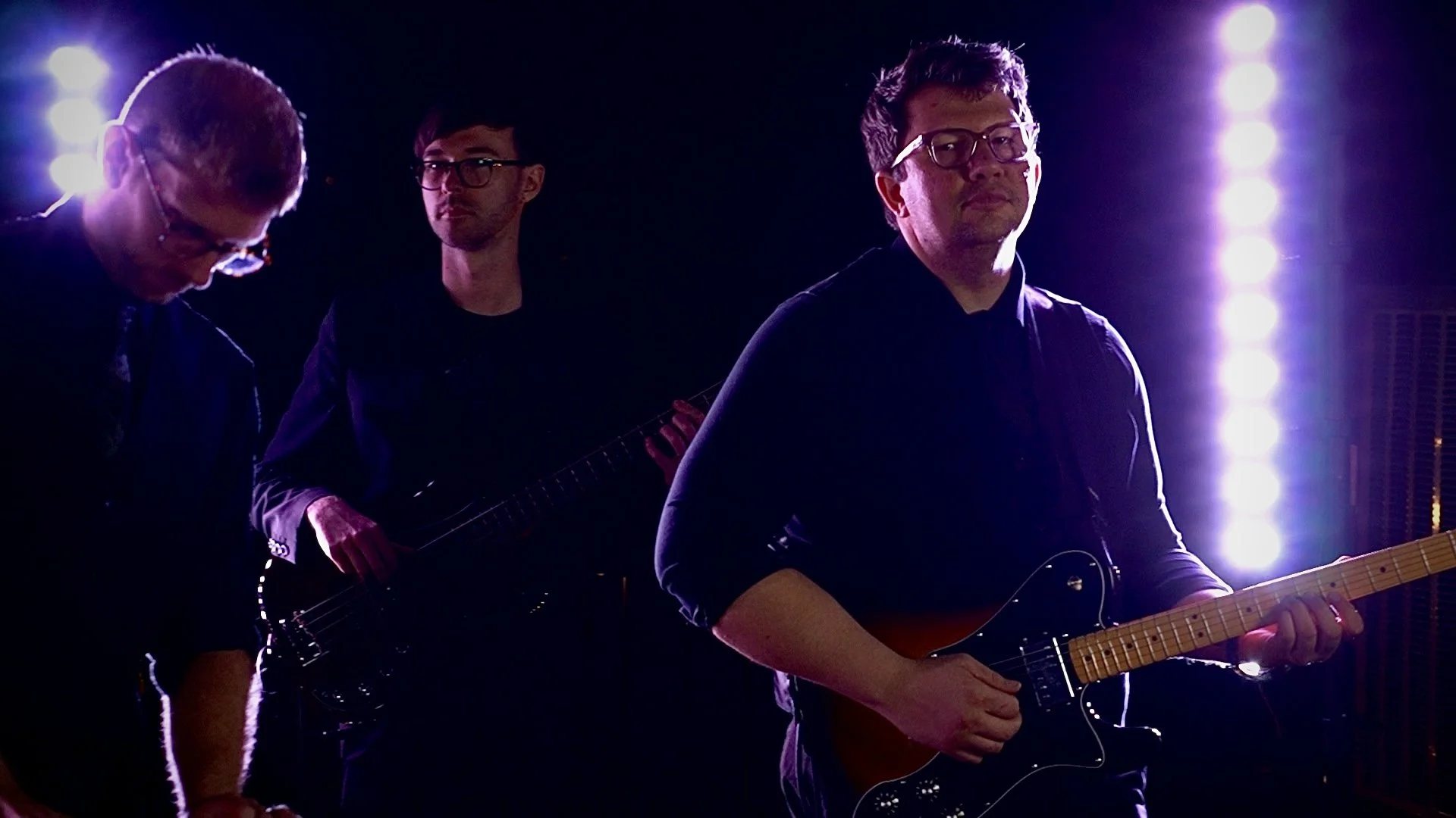 Three men playing guitars on stage with bright purple stage lights in the background.