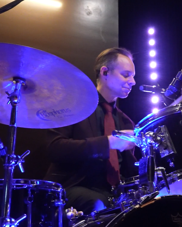 A drummer playing a drum kit on stage, illuminated by blue and purple stage lights.