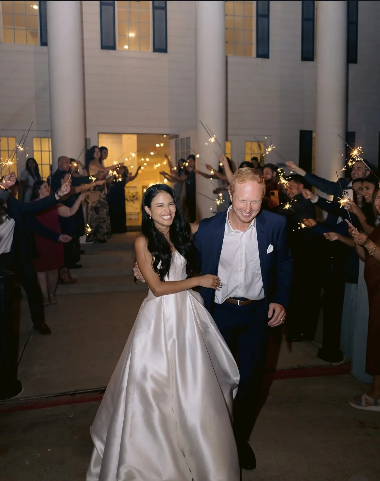 A newlywed couple in wedding attire walking out of a building, smiling, while guests hold sparklers on either side celebrating.