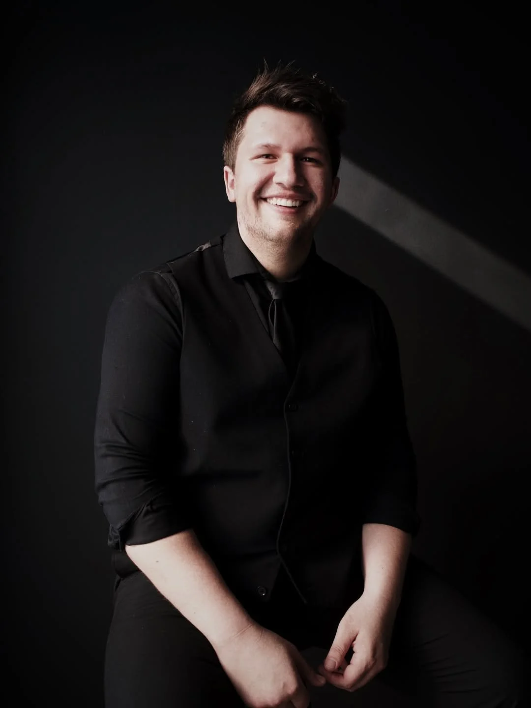 A young man with short, dark, messy hair smiling, wearing a black, long-sleeved button-up shirt, sitting against a dark background with a light shining on his face.